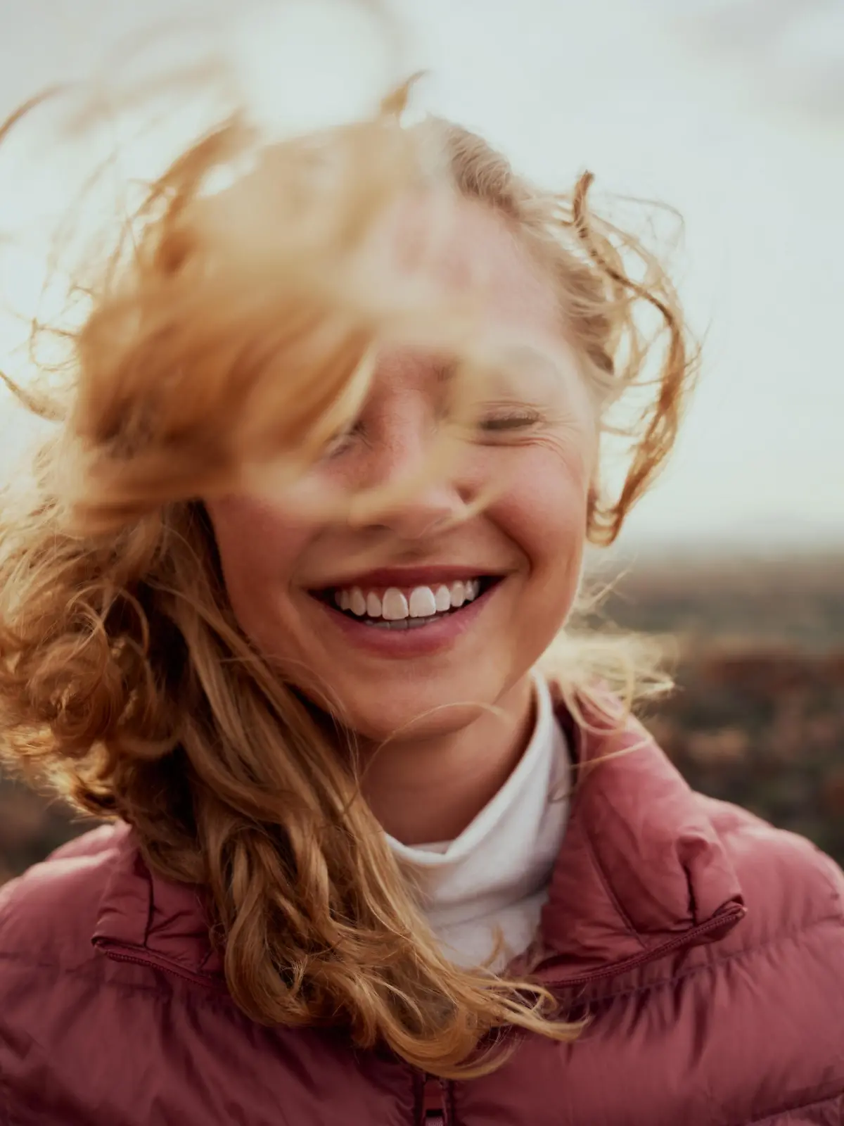 Portrait of young smiling woman face partially covered with flying hair standing at mountain - carefree woman