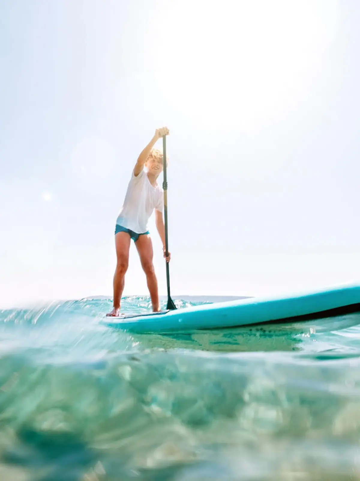 Under the water view angle to the smiling blonde teenager boy rowing stand up paddle board. Active family summer vacation time near the sea concept image.