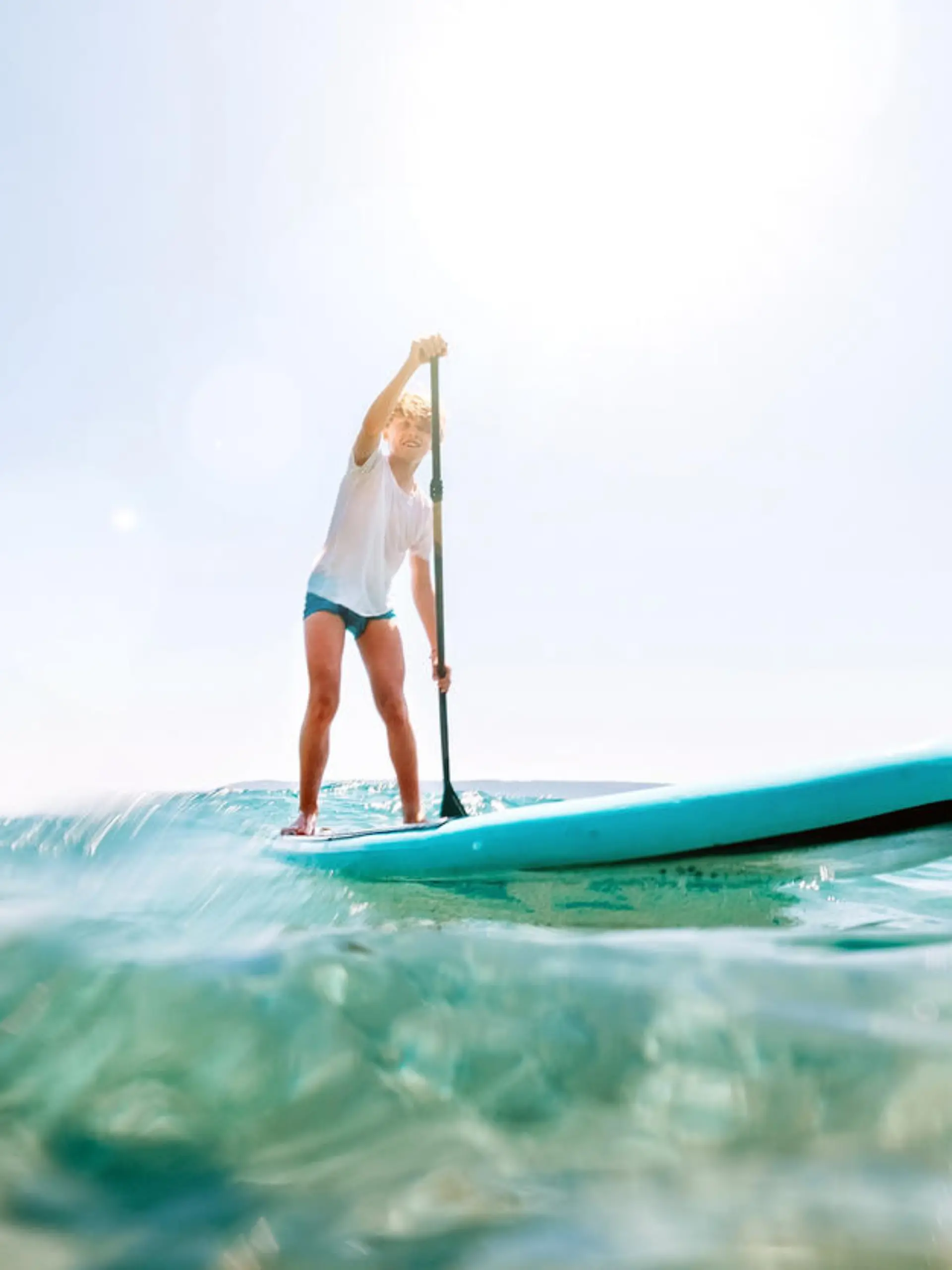 Under the water view angle to the smiling blonde teenager boy rowing stand up paddle board. Active family summer vacation time near the sea concept image.
