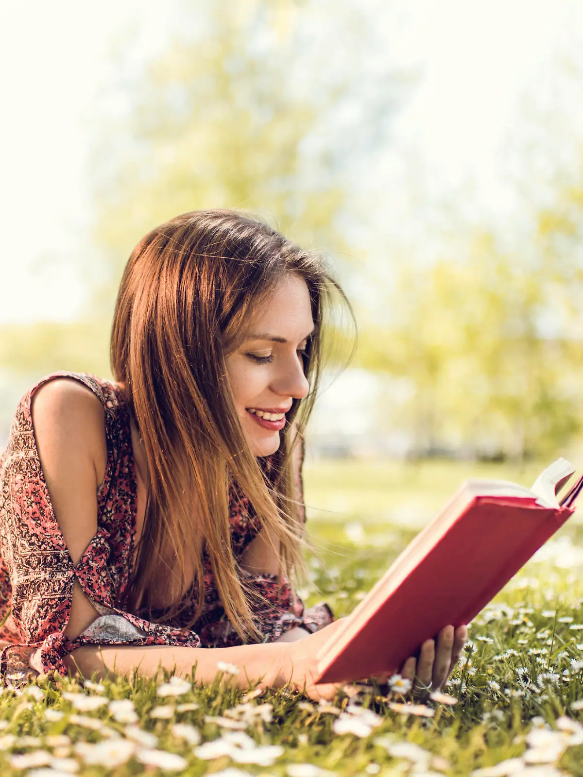 Young happy female student enjoying in spring day at the park and learning from a book.