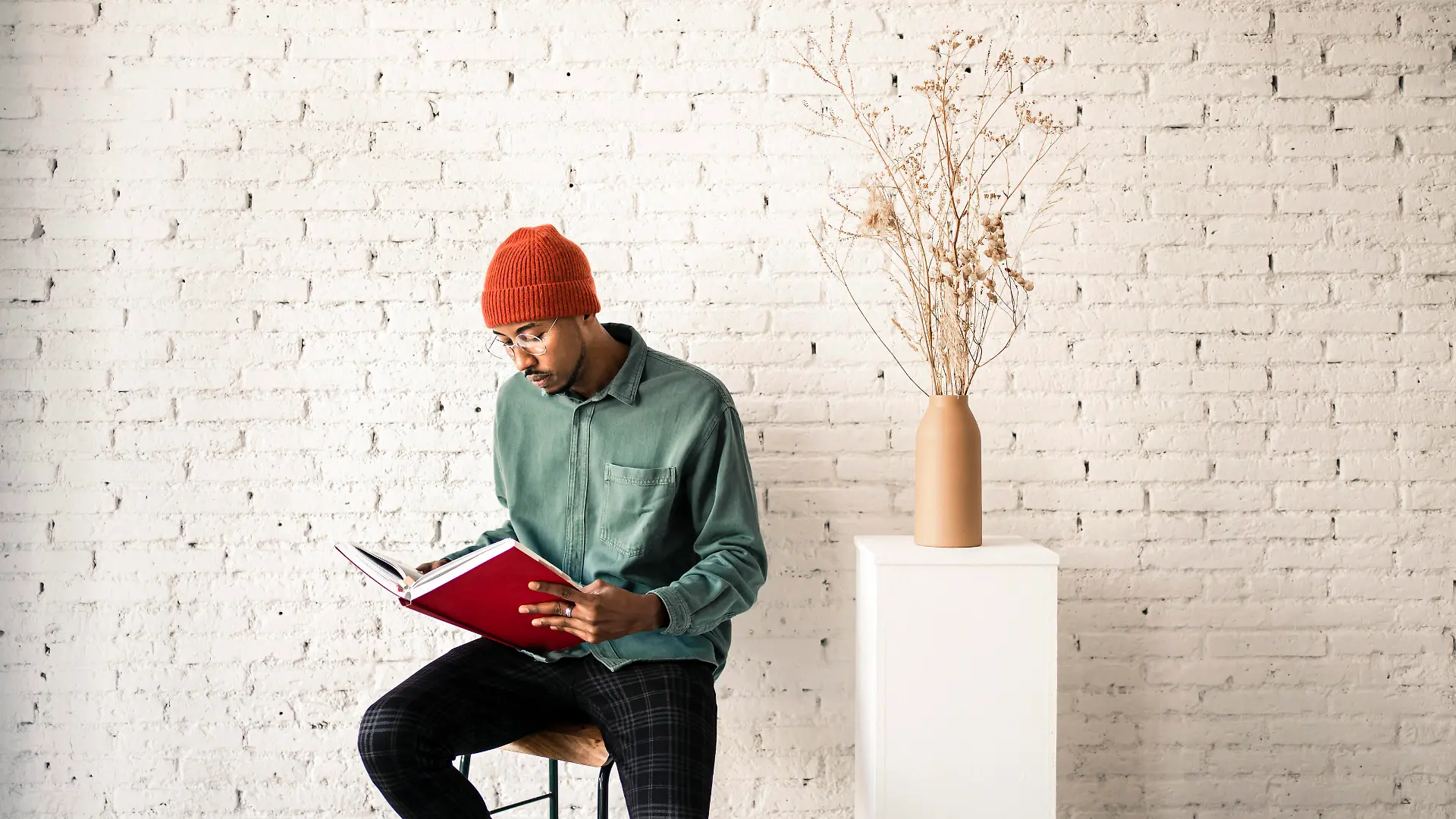 Man in knit hat reading book by dried plant vase against white brick wall model released Symbolfoto property released RCPF00507