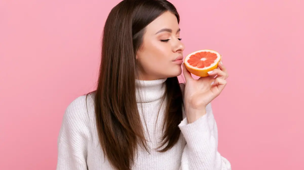 Beautiful female smelling half slice grapefruit, enjoying tasty vitamin fruits, healthy nutrition, wearing white casual style sweater. Indoor studio shot isolated on pink background.