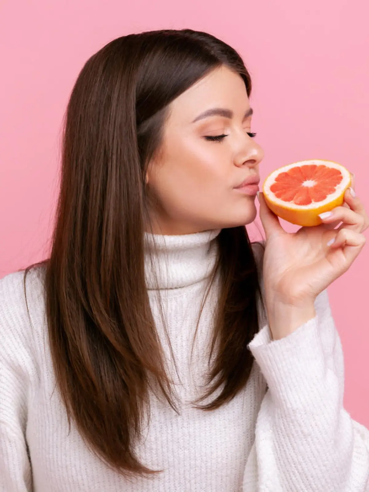 Beautiful female smelling half slice grapefruit, enjoying tasty vitamin fruits, healthy nutrition, wearing white casual style sweater. Indoor studio shot isolated on pink background.