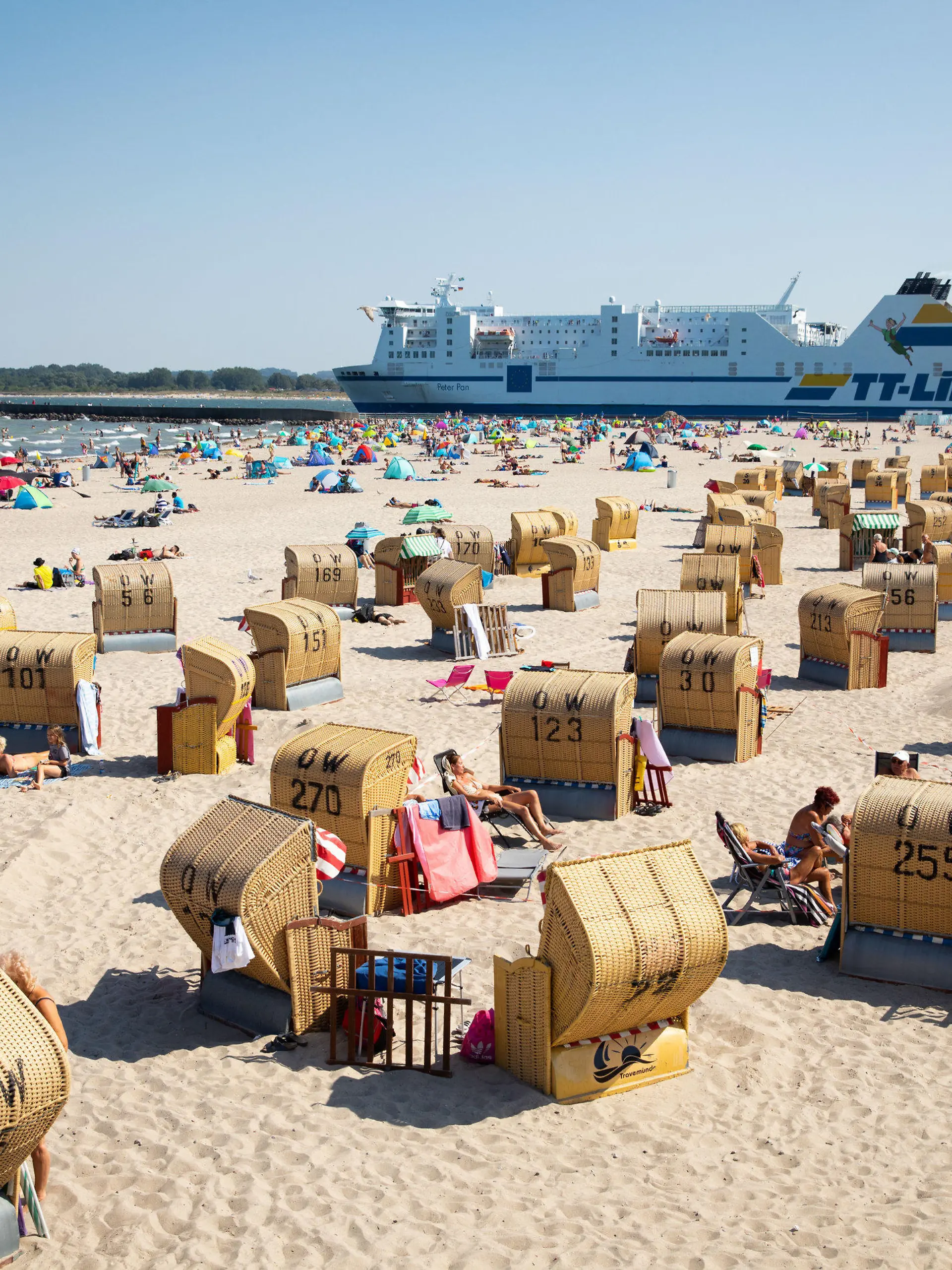 ARCHIV - 11.08.2020, Schleswig-Holstein, Travemünde: Urlauber und Tagesgäste verbringen bei strahlendem Sonnenschein und warmen Temperaturen den Tag am Strand von Travemünde, während im Hintergrund eine Fähre der TT-Line ausläuft. Der «Corona-Sommer» 2020 hat in vielen Tourismusorten in Schleswig-Holstein einen weniger großen Schaden angerichtet, als zunächst befürchtet. Foto: Christian Charisius/dpa - ACHTUNG: Verwendung nur im vollen Format +++ dpa-Bildfunk +++