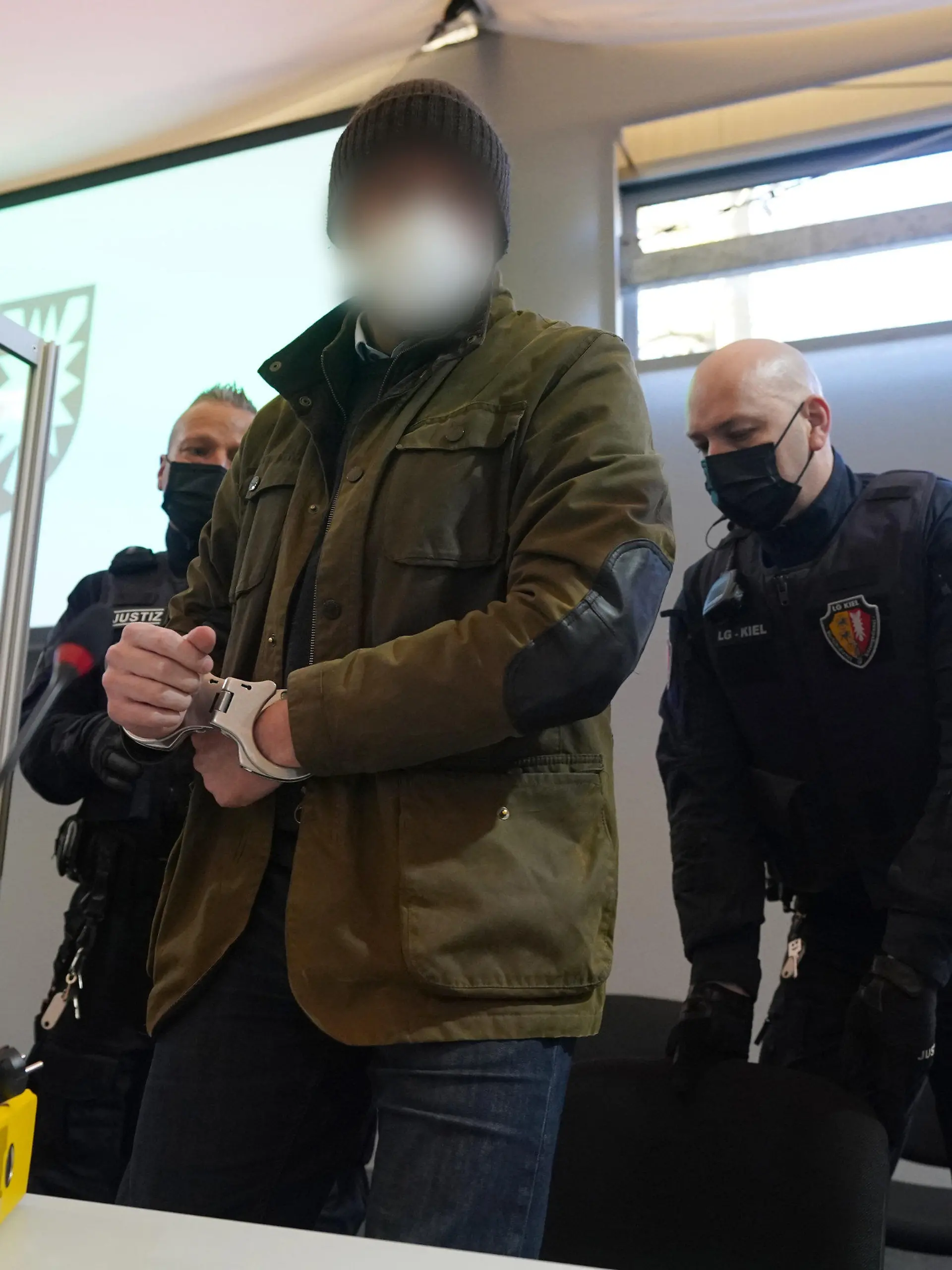 07.03.2022, Schleswig-Holstein, Kiel: Justizbeamte bringen den Angeklagten (M) in Handschellen in den Sitzungssaal der Außenstelle des Landgerichts Kiel zu Beginn des Prozesstages wegen eines Dreifachmordes. Am 19. Mai 2021 soll der Mann zuerst seine von ihm getrennt lebende Ehefrau und deren neuen Bekannten in Dänischenhagen mit einer Maschinenpistole erschossen und danach in Kiel einen gemeinsamen Bekannten des Ehepaares mit einer halbautomatischen Pistole getötet haben. Mit der zweiten Waffe stellte er sich am Abend in Hamburg der Polizei. Foto: Marcus Brandt/dpa - ACHTUNG: Der Angeklagte wurde aus rechtlichen Gründen gepixelt +++ dpa-Bildfunk +++