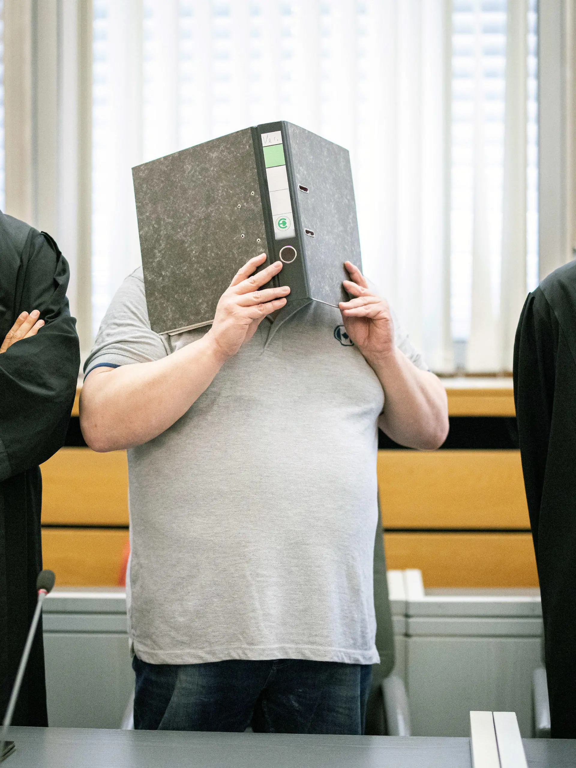 12.08.2020, Nordrhein-Westfalen, Essen: Der Angeklagte (M) steht zu Beginn des Prozesses im Gerichtssaal des Landgerichts mit einem Aktenordner vor dem Gesicht zwischen seinen Anwälten Stefan Tierel (l) und Leonhardt Mühlenfeld. Der 47 Jahre alte Mann aus Krefeld soll eine Bekannte im Juni 2019 mit einer über den Kopf gezogenen Plastiktüte erstickt haben. Hintergrund der Tat soll Eifersucht gewesen sein. Angeblich wollte sich die Frau von ihm trennen. Die Leiche ist bis heute nicht gefunden worden. Foto: Marcel Kusch/dpa - ACHTUNG: Schrift auf Ordnerrücken wurde aus rechtlichen Gründen gepixelt +++ dpa-Bildfunk +++