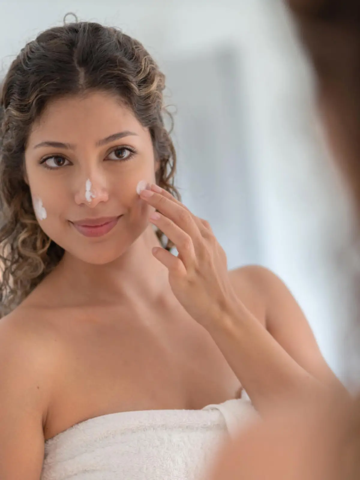 Portrait of a beautiful woman in the bathroom applying moisturizer on her face and looking in the mirror