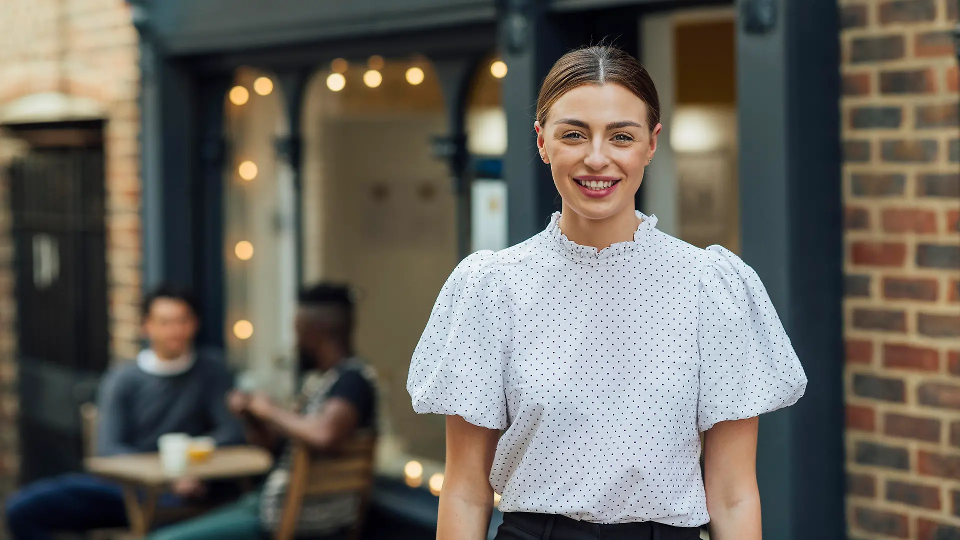 Waist up portrait of a young beautiful woman dressed formally with her hair tied back standing outside of a cafe looking at the camera. There are students out of focus sitting at an outdoor cafe table behind her. She is very cheerful looking at the camera.