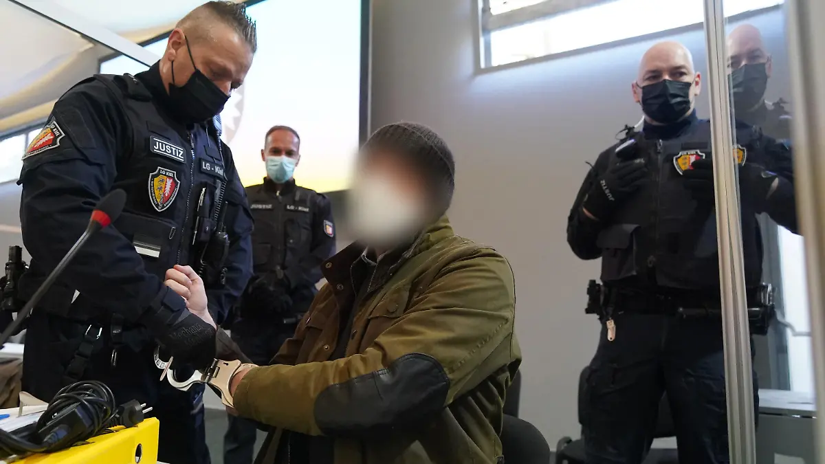 07.03.2022, Schleswig-Holstein, Kiel: Ein Justizbeamter entfernt die Handschellen des Angeklagten (2.v.r) im Sitzungssaal der Außenstelle des Landgerichts Kiel zu Beginn des Prozesstages wegen eines Dreifachmordes. Am 19. Mai 2021 soll der Mann zuerst seine von ihm getrennt lebende Ehefrau und deren neuen Bekannten in Dänischenhagen mit einer Maschinenpistole erschossen und danach in Kiel einen gemeinsamen Bekannten des Ehepaares mit einer halbautomatischen Pistole getötet haben. Mit der zweiten Waffe stellte er sich am Abend in Hamburg der Polizei. Foto: Marcus Brandt/dpa - ACHTUNG: Der Angeklagte wurde aus rechtlichen Gründen gepixelt +++ dpa-Bildfunk +++