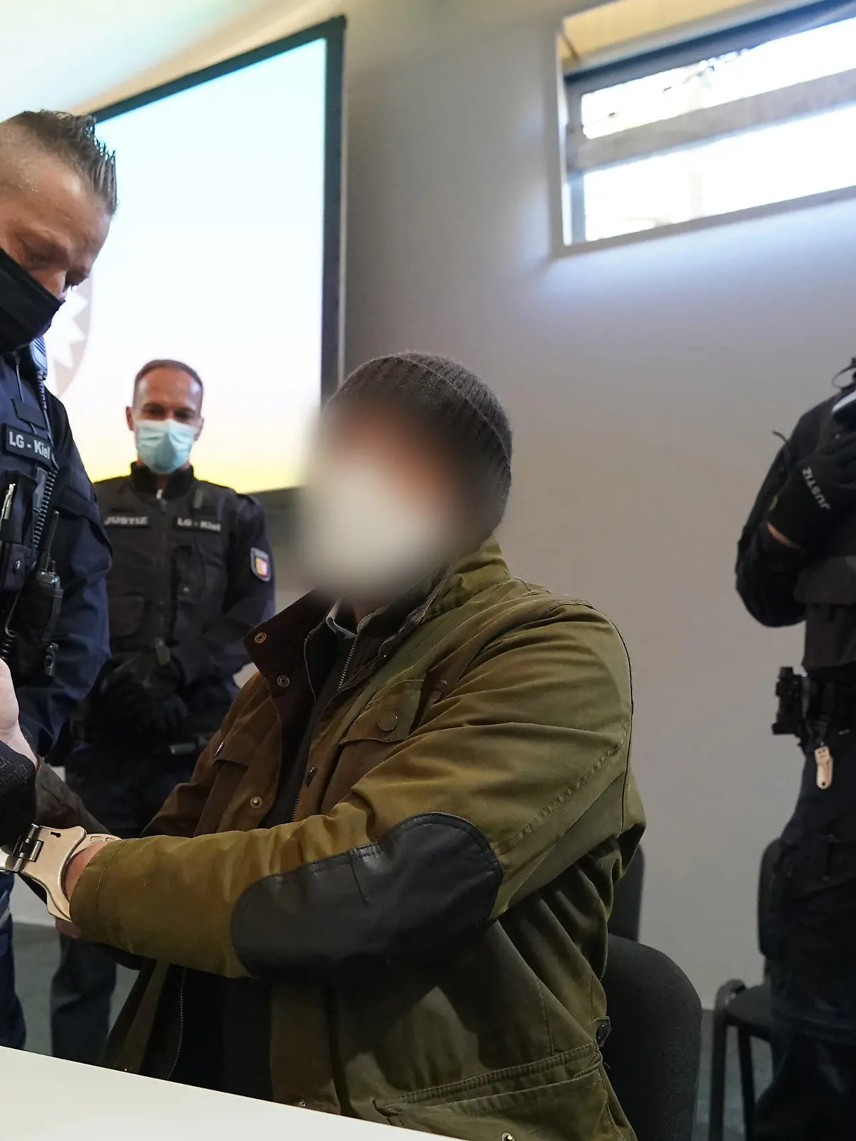 07.03.2022, Schleswig-Holstein, Kiel: Ein Justizbeamter entfernt die Handschellen des Angeklagten (2.v.r) im Sitzungssaal der Außenstelle des Landgerichts Kiel zu Beginn des Prozesstages wegen eines Dreifachmordes. Am 19. Mai 2021 soll der Mann zuerst seine von ihm getrennt lebende Ehefrau und deren neuen Bekannten in Dänischenhagen mit einer Maschinenpistole erschossen und danach in Kiel einen gemeinsamen Bekannten des Ehepaares mit einer halbautomatischen Pistole getötet haben. Mit der zweiten Waffe stellte er sich am Abend in Hamburg der Polizei. Foto: Marcus Brandt/dpa - ACHTUNG: Der Angeklagte wurde aus rechtlichen Gründen gepixelt +++ dpa-Bildfunk +++