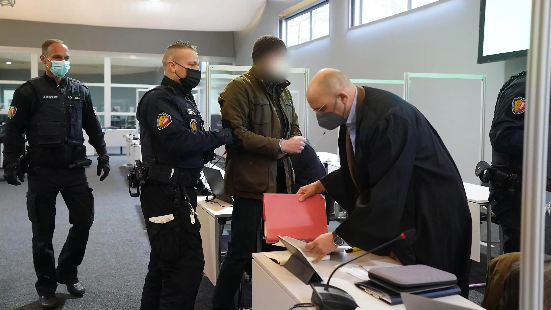 07.03.2022, Schleswig-Holstein, Kiel: Justizbeamte bringen den Angeklagten (3.v.l.) in Handschellen in den Sitzungssaal der Außenstelle des Landgerichts Kiel zu Beginn des Prozesstages wegen eines Dreifachmordes. Vorne rechts im Bild steht sein Verteidiger, Abdou Gabbar. Am 19. Mai 2021 soll der Mann zuerst seine von ihm getrennt lebende Ehefrau und deren neuen Bekannten in Dänischenhagen mit einer Maschinenpistole erschossen und danach in Kiel einen gemeinsamen Bekannten des Ehepaares mit einer halbautomatischen Pistole getötet haben. Mit der zweiten Waffe stellte er sich am Abend in Hamburg der Polizei. Foto: Marcus Brandt/dpa - ACHTUNG: Der Angeklagte wurde aus rechtlichen Gründen gepixelt +++ dpa-Bildfunk +++