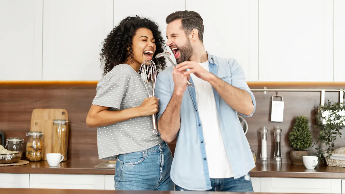 Happy interracial couple dancing in the kitchen, singing while cooking breakfast or dinner, young African American woman and a handsome man smiling and having fun together, holding kitchen utensils