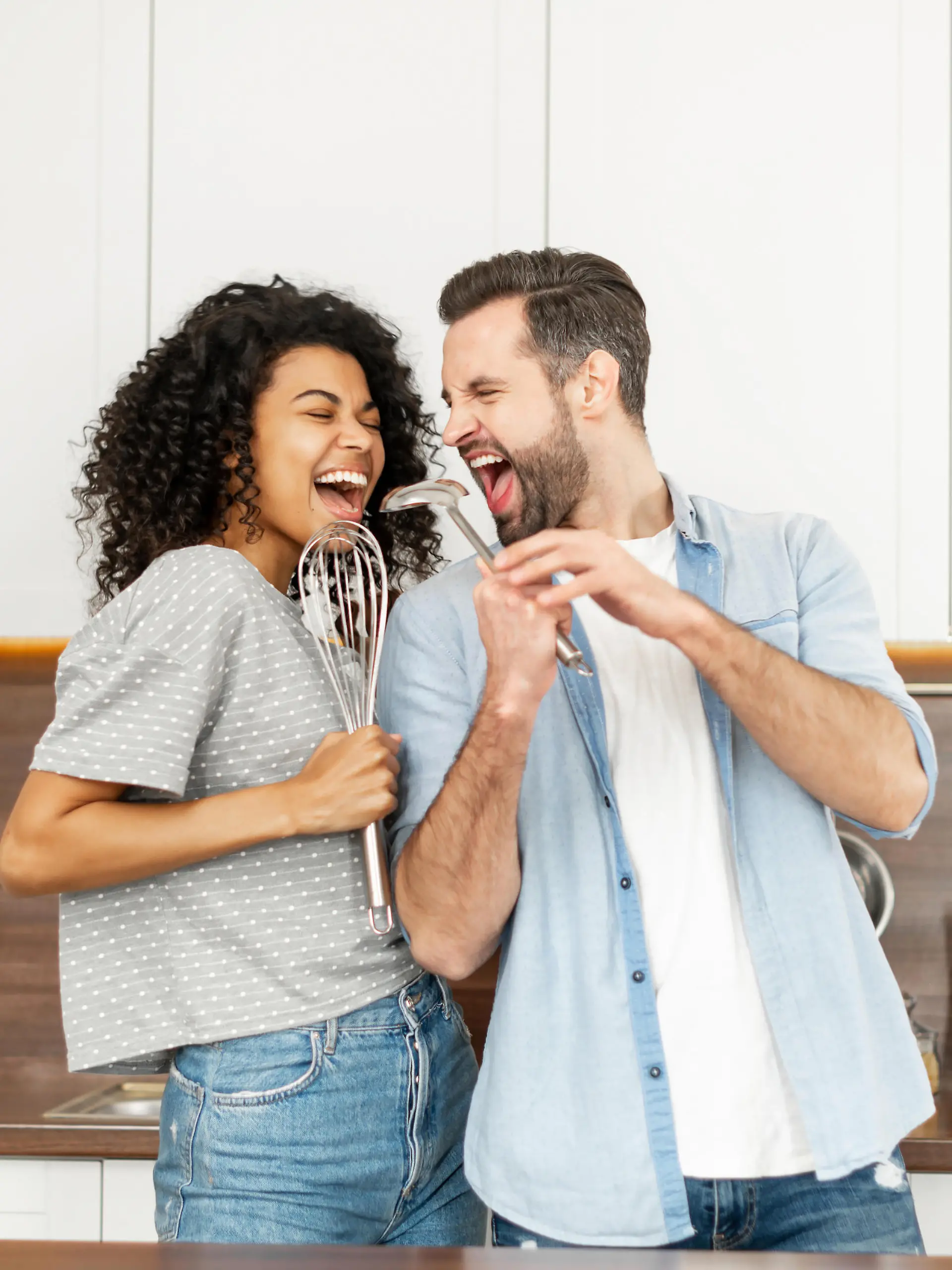 Happy interracial couple dancing in the kitchen, singing while cooking breakfast or dinner, young African American woman and a handsome man smiling and having fun together, holding kitchen utensils
