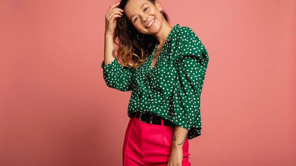 Young woman dressed in a green polka dot shirt standing in front of a pink background.