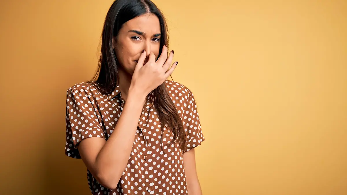 Young beautiful brunette woman wearing casual shirt over isolated yellow background smelling something stinky and disgusting, intolerable smell, holding breath with fingers on nose. Bad smell