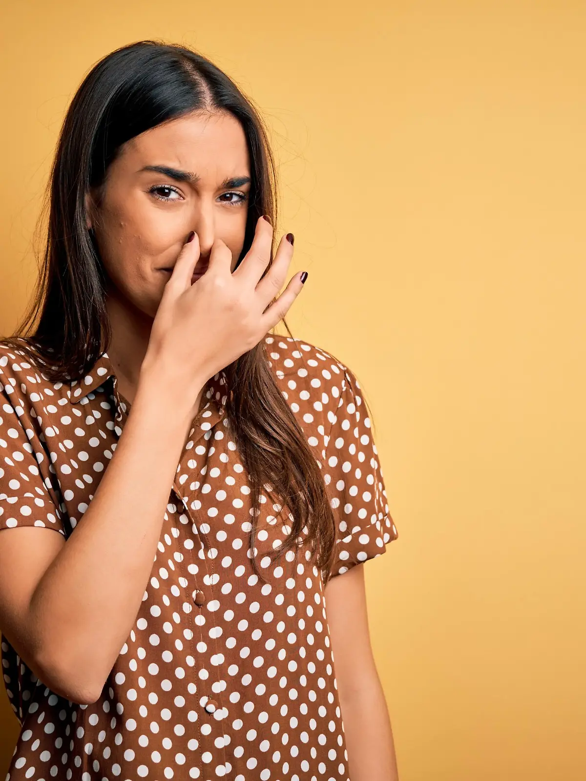 Young beautiful brunette woman wearing casual shirt over isolated yellow background smelling something stinky and disgusting, intolerable smell, holding breath with fingers on nose. Bad smell