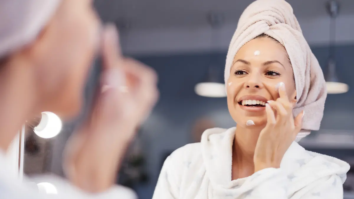 Portrait of beautiful Caucasian woman in towel looking herself in the mirror and applying face cream. She is going through morning beauty routine in bathroom. Skin Care and Hygiene