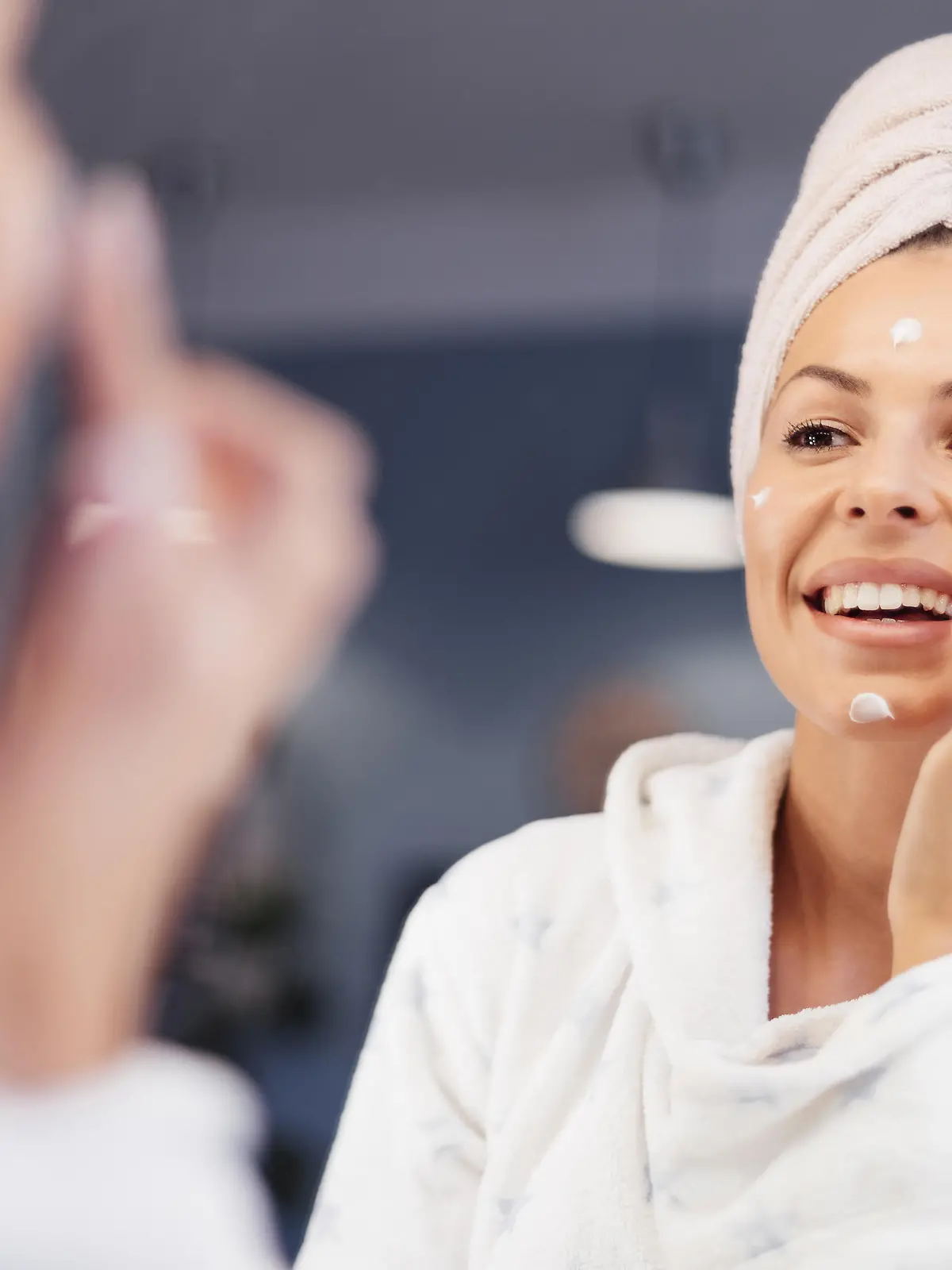 Portrait of beautiful Caucasian woman in towel looking herself in the mirror and  applying face cream. She is going through morning beauty routine in bathroom. Skin Care and Hygiene