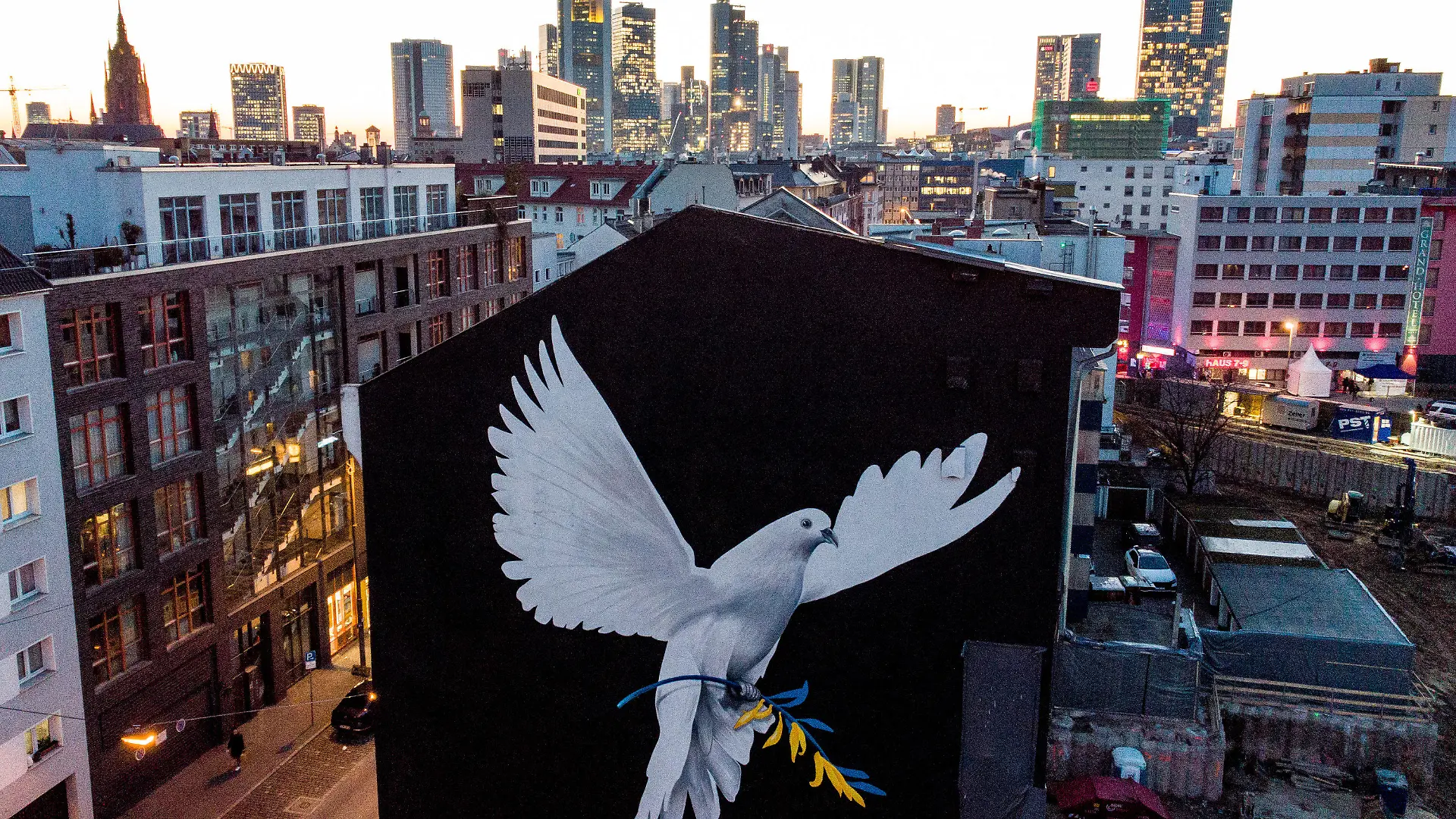 28.02.2022, Hessen, Frankfurt/Main: Eine riesige Friedenstaube hat der Künstler Justus Becker an eine Hausfassade vor der Bankenskyline von Frankfurt am Main gemalt. Die Taube trägt einen Ölzweig in den ukrainischen Nationalfarben blau und gelb. Mit dem Wandgemälde an der Fassade eines fünfgeschossigen Hauses hat der Frankfurter Künstler J. Becker ein Zeichen angesichts des Krieges in der Ukraine gesetzt. Foto: Boris Roessler/dpa - ACHTUNG: Verwendung nur im vollen Format und nur zur redaktionellen Verwendung +++ dpa-Bildfunk +++