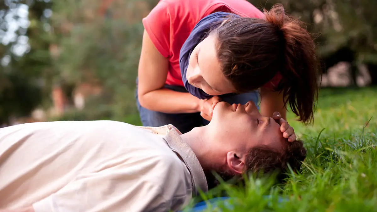 First aid - Look, listen and feel for breathing. Woman is performing first and on a man laying in the grass. She is pulling his head back, listening to his breath and looking at his chest to check if he is breathing. The man is laying unconscious. 