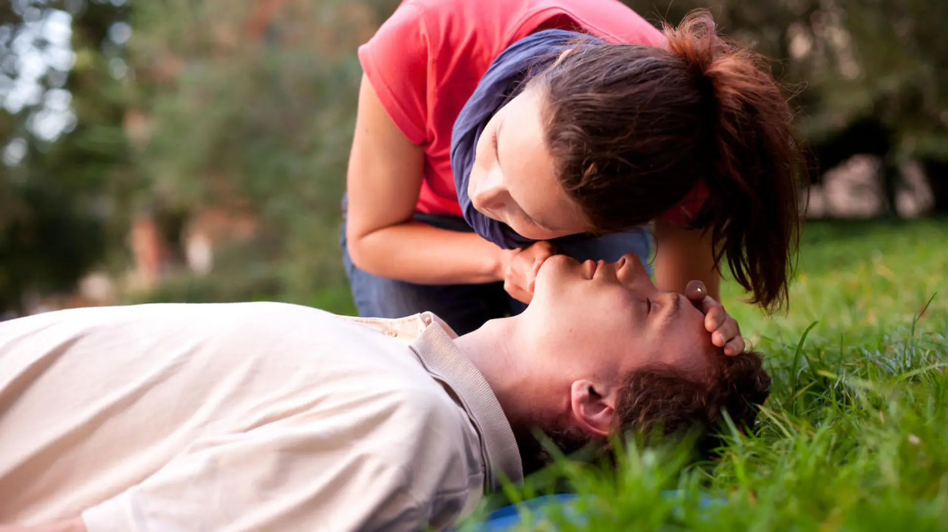 First aid - Look, listen and feel for breathing. Woman is performing first and on a man laying in the grass. She is pulling his head back, listening to his breath and looking at his chest to check if he is breathing. The man is laying unconscious.