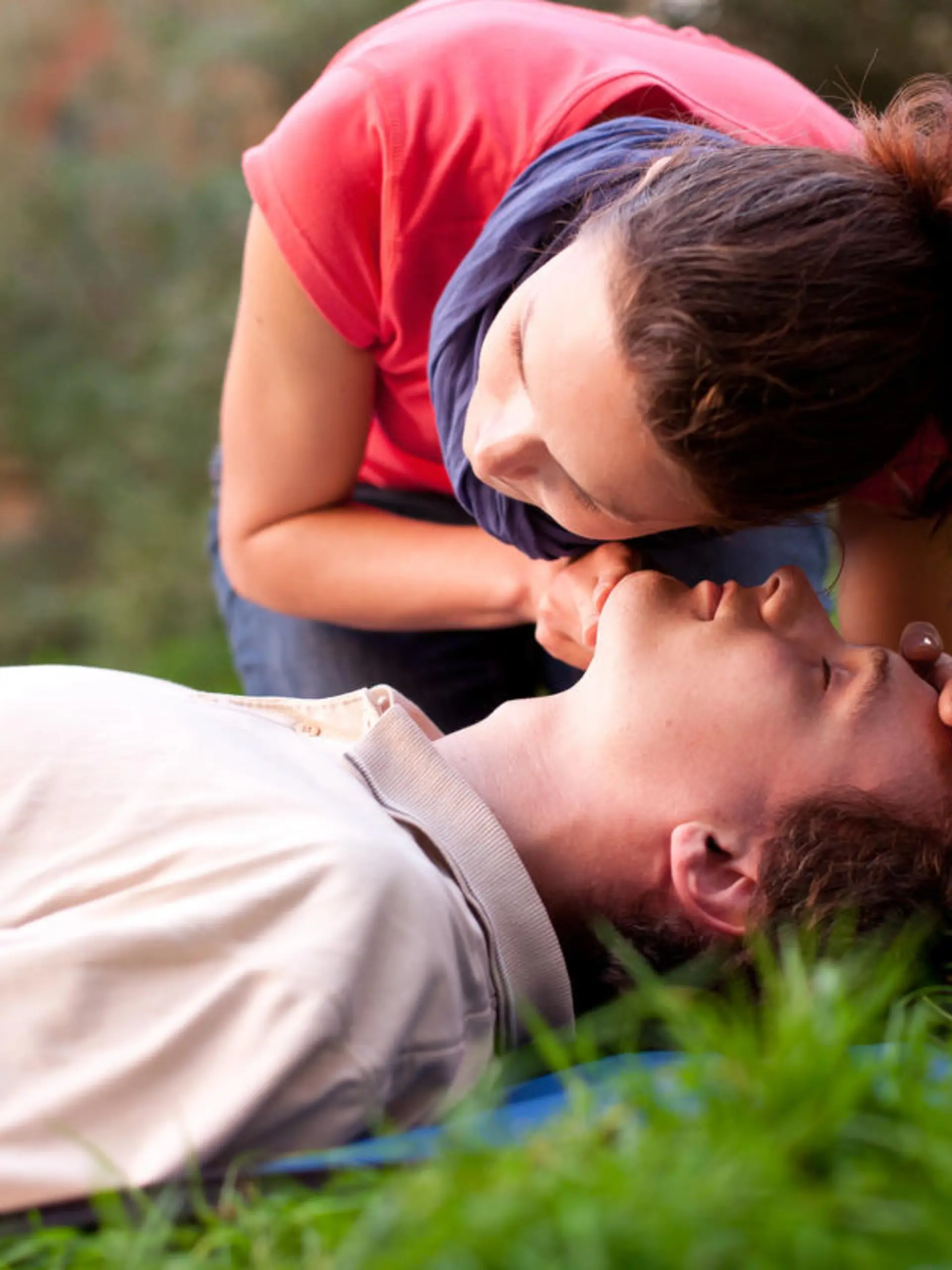 First aid - Look, listen and feel for breathing. Woman is performing first and on a man laying in the grass. She is pulling his head back, listening to his breath and looking at his chest to check if he is breathing. The man is laying unconscious. 