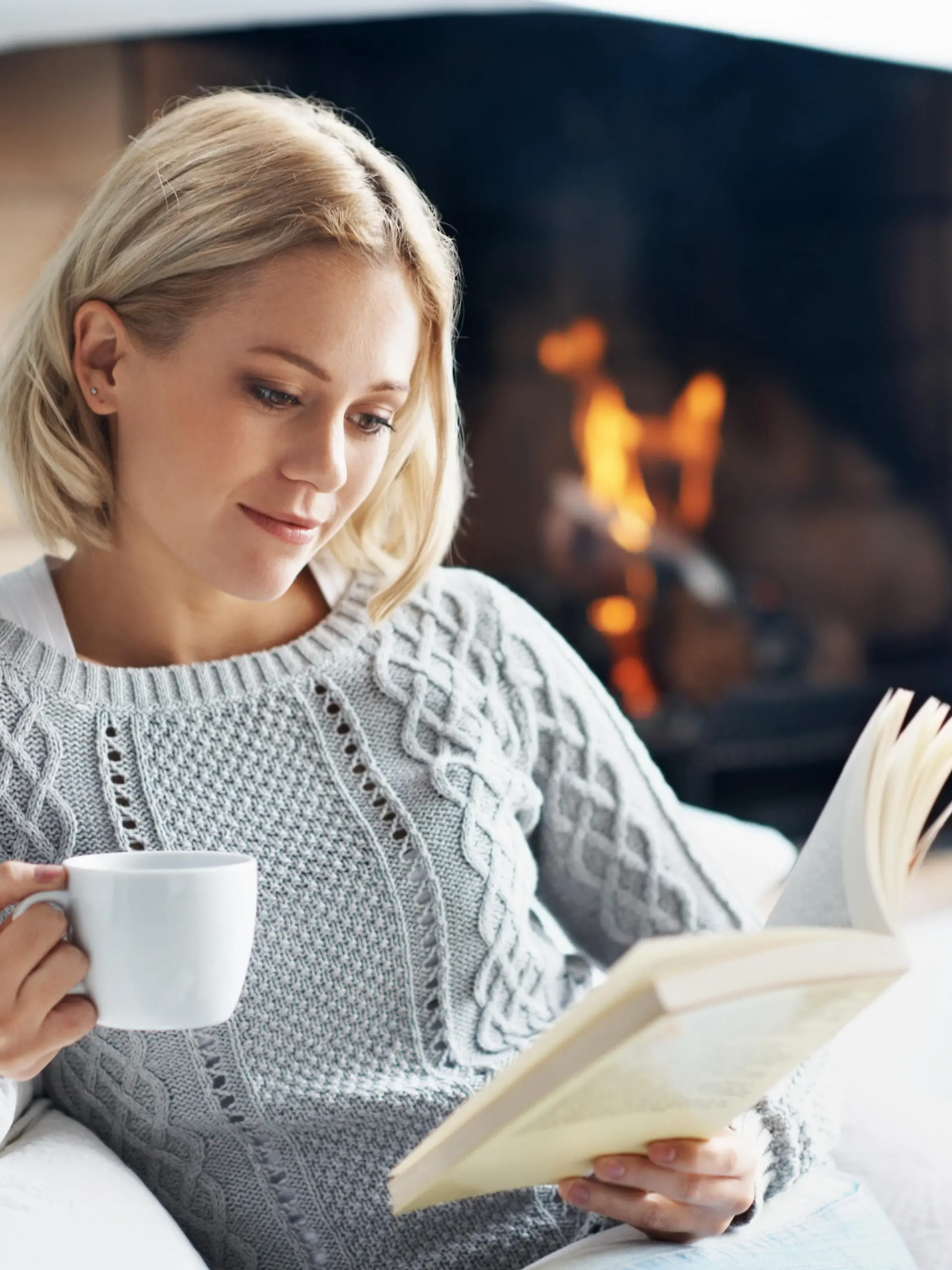 A beautiful young woman reading a book and enjoying a warm beverage near a fireplace