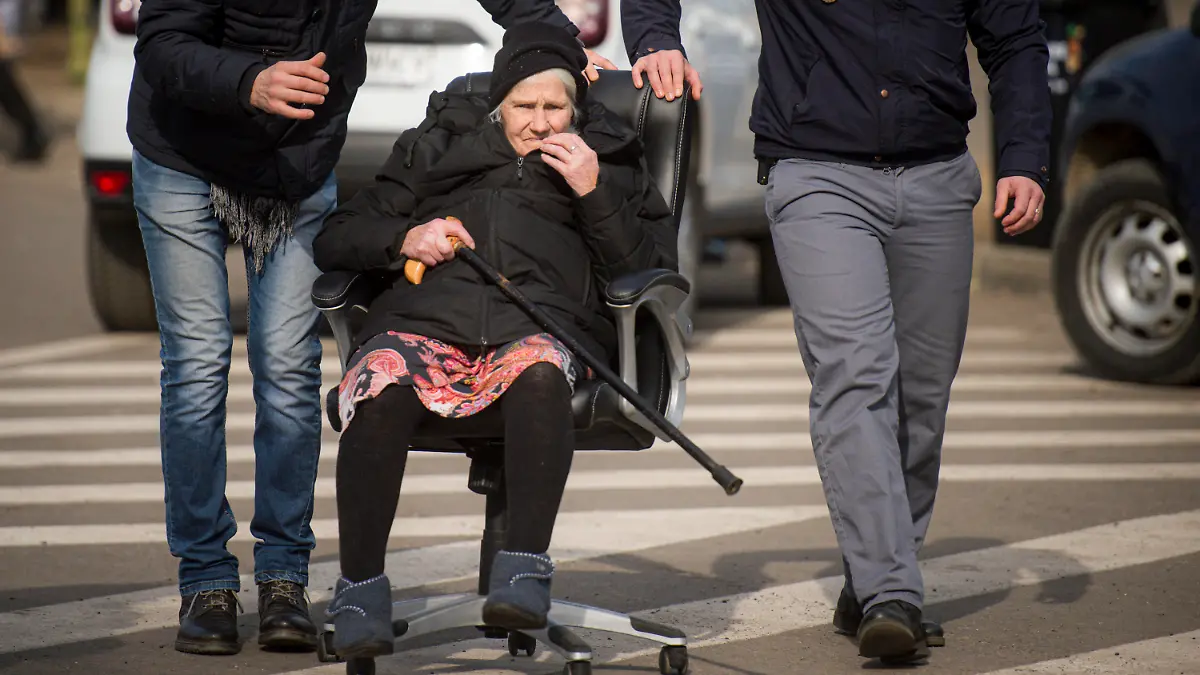 27.02.2022, Rumänien, Siret: Grenzpolizisten schieben eine ältere Dame in einem Bürostuhl an der ukrainischen-rumänischen Grenze. Wegen des russischen Angriffs auf die Ukraine sind nach Angaben des UN-Flüchtlingshilfswerks (UNHCR) etwa 368 000 Menschen auf der Flucht. Foto: Alexandru Dobre/AP/dpa +++ dpa-Bildfunk +++