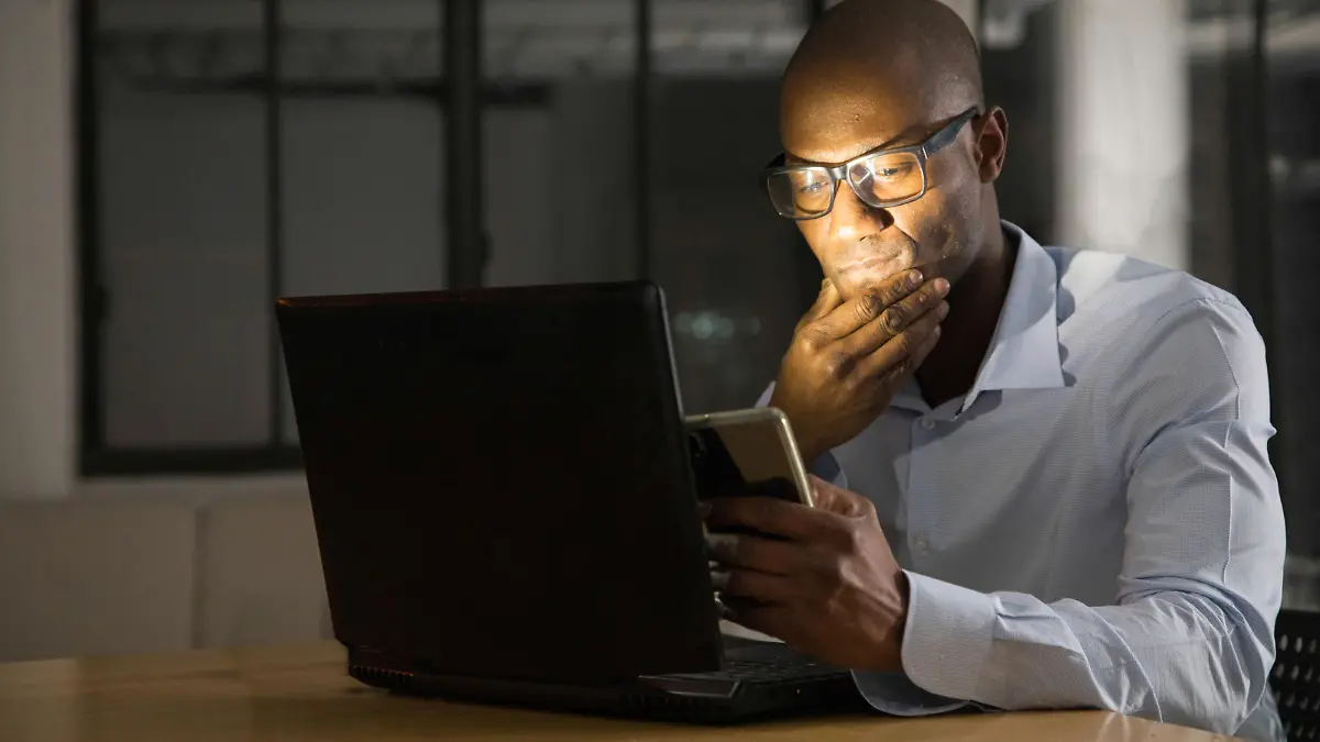 Mature black man while working on his laptop and mobile phone at night. He looks pensively at what he sees on his screens.
