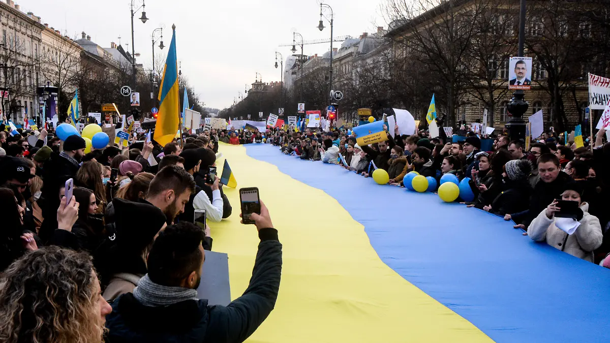 26.02.2022, Ungarn, Budapest: Demonstranten stehen um eine große ukrainische Flagge versammelt und protestieren gegen den russischen Einmarsch in die Ukraine. Russische Truppen haben den erwarteten Angriff auf die Ukraine gestartet und drangen in die Hauptstadt vor. Foto: Justin Spike/AP/dpa +++ dpa-Bildfunk +++