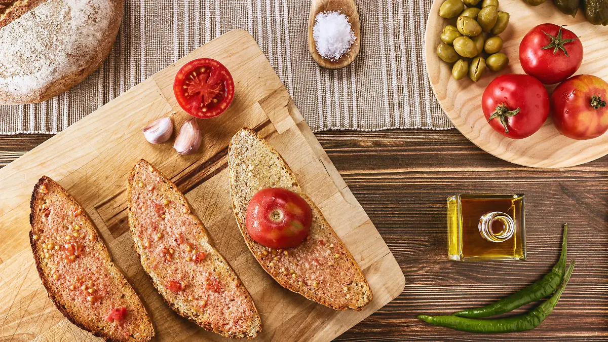 Two slices of bread with tomato pulp and olive oil , one slice with half ramillet tomato and the rest of the ingredients like garlic, typical "ramillet" tomatoes from Mallorca, oil bottle, green olives and pickles. All distributed on a dark brown wooden table.