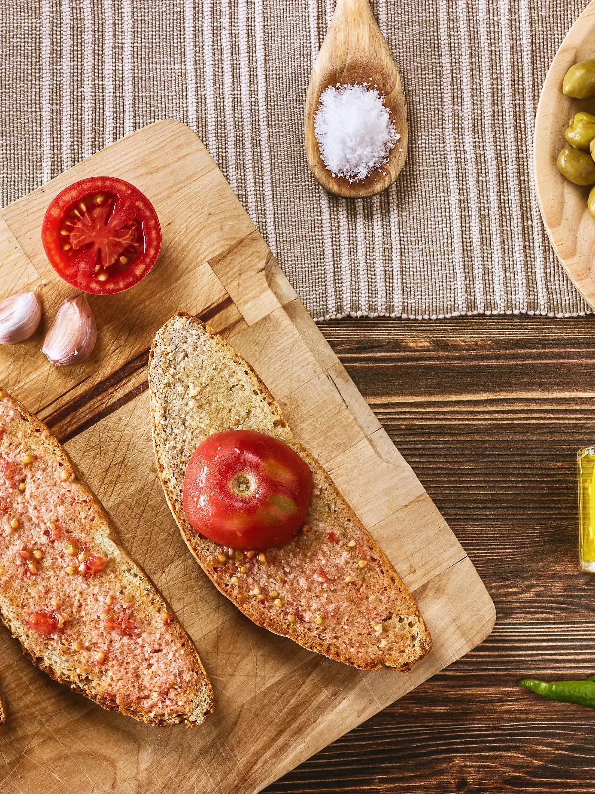 Two slices of bread with tomato pulp and olive oil , one slice with half ramillet tomato and the rest of the ingredients like garlic, typical "ramillet" tomatoes from Mallorca, oil bottle, green olives and pickles. All distributed on a dark brown wooden table.