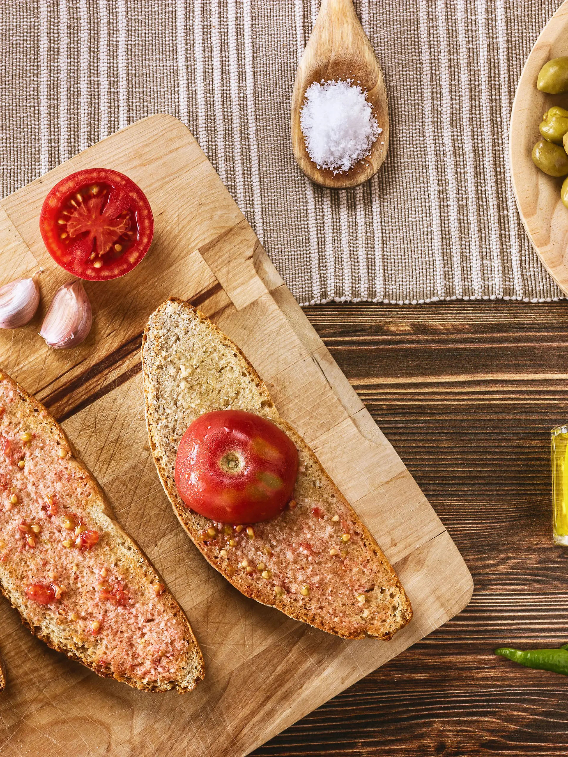Two slices of bread with tomato pulp and olive oil , one slice with half ramillet tomato and the rest of the ingredients like garlic, typical "ramillet" tomatoes from Mallorca, oil bottle, green olives and pickles. All distributed on a dark brown wooden table.