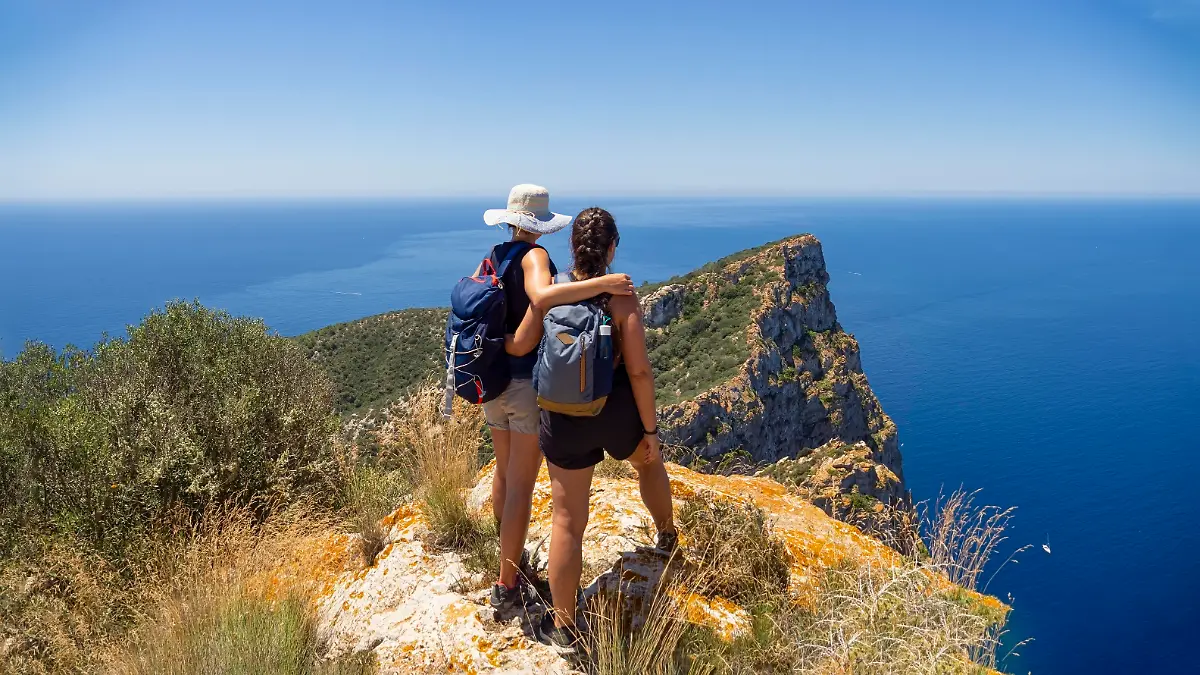 Two women hikers on mountain summit overlooking the mediterranean sea. Outdoor adventure day trek with friends. Aspiration, exploration and inspiring lifestyle concept. Panoramic from a mountain peak.