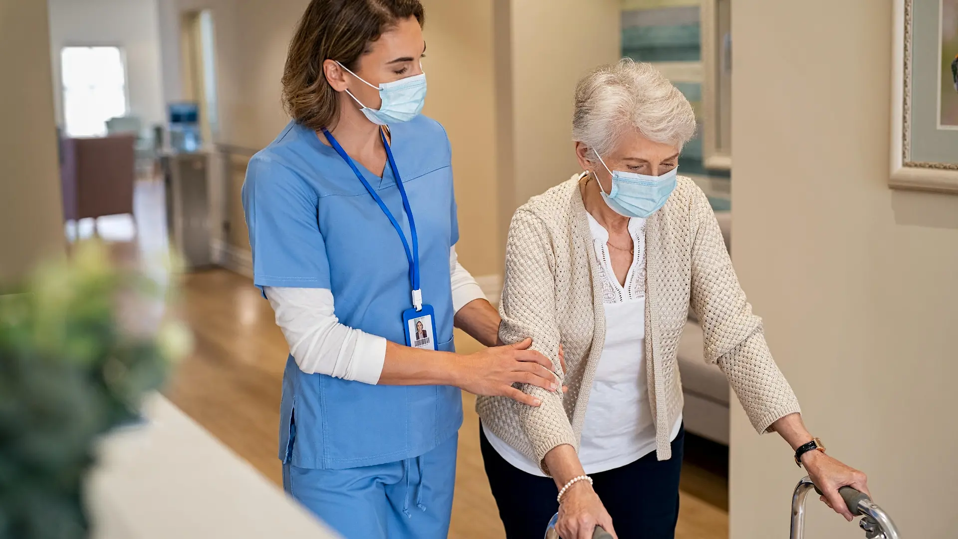 Smiling nurse with face mask helping senior woman to walk around the nursing home with walker. Young lovely nurse helping old woman with surgical mask for safety against covid-19 using a walking frame in hospital corridor. Friendly caregiver and elderly patient walking in hallway wearing protective face mask during covid pandemic.