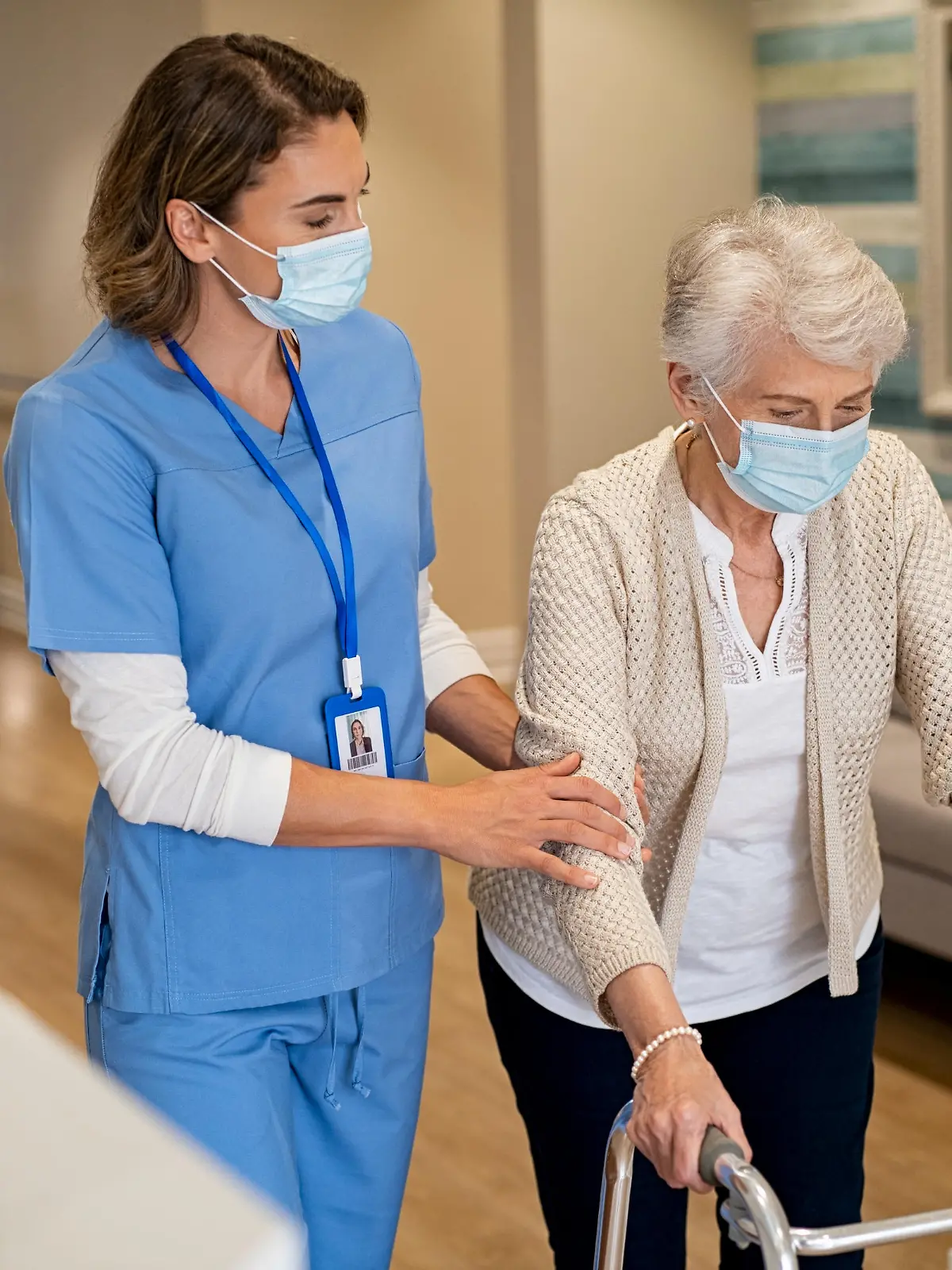 Smiling nurse with face mask helping senior woman to walk around the nursing home with walker. Young lovely nurse helping old woman with surgical mask for safety against covid-19 using a walking frame in hospital corridor. Friendly caregiver and elderly patient walking in hallway wearing protective face mask during covid pandemic.