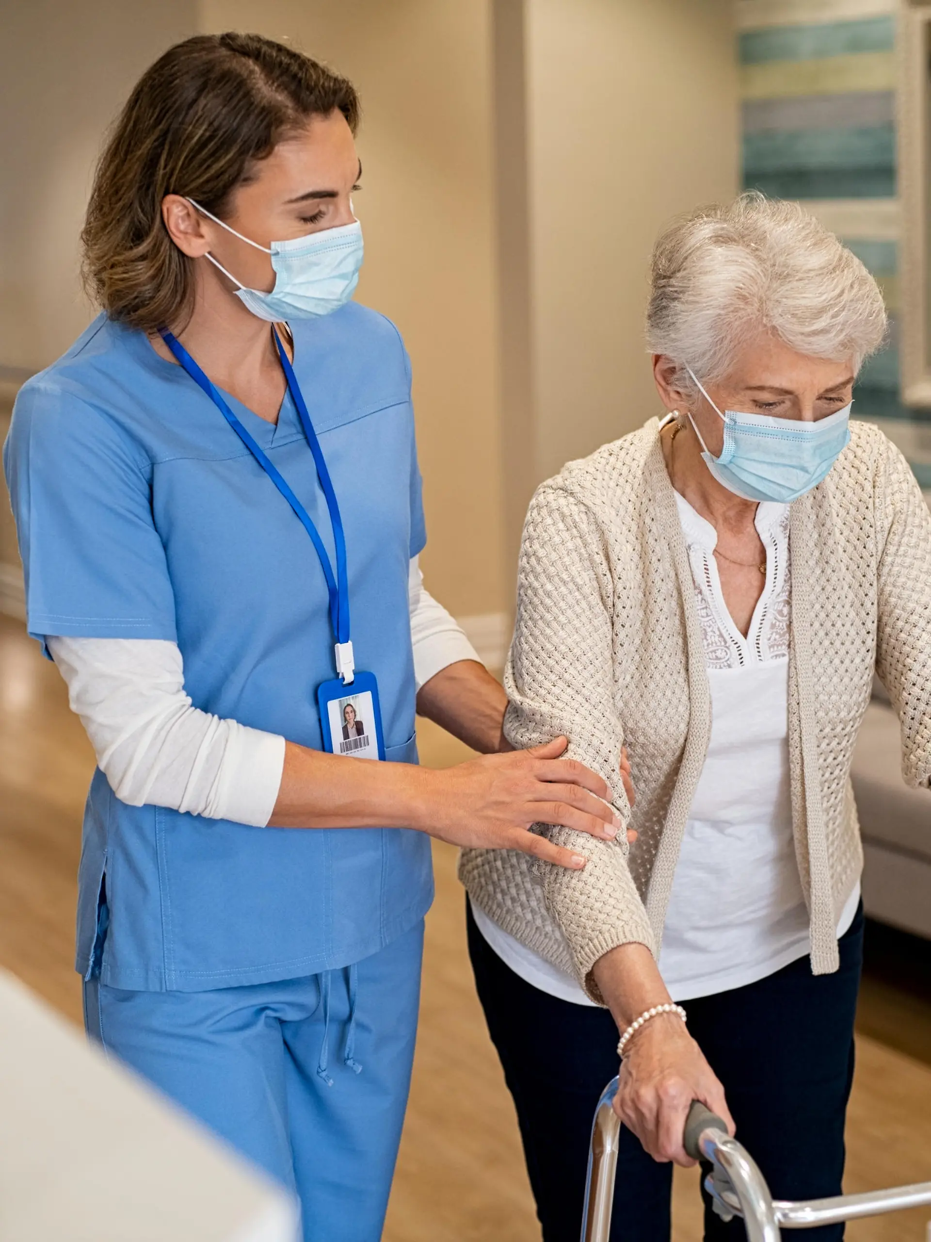 Smiling nurse with face mask helping senior woman to walk around the nursing home with walker. Young lovely nurse helping old woman with surgical mask for safety against covid-19 using a walking frame in hospital corridor. Friendly caregiver and elderly patient walking in hallway wearing protective face mask during covid pandemic.