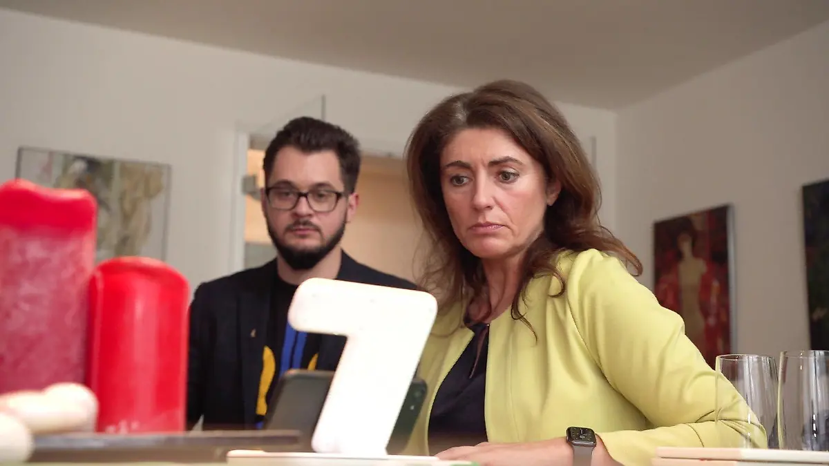 Vilen Adamskyi und Linda Mai sitzen an einem Tisch mit einer hell-orangenen Tischdecke und schauen mit ernstem Gesicht nach unten. Im Vordergrund des Bild sieht man einen Blumenstrauß von gelben Tulpen.