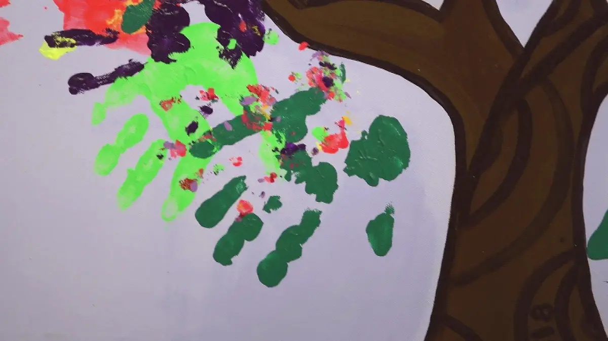 Zwei grüne Kinder-Handabdrücke werden im Close-Up gezeigt. Sie sind Teil eines Gemäldes, in dem viele bunte Kinderhandabdrücke eine Baumkrone bilden.