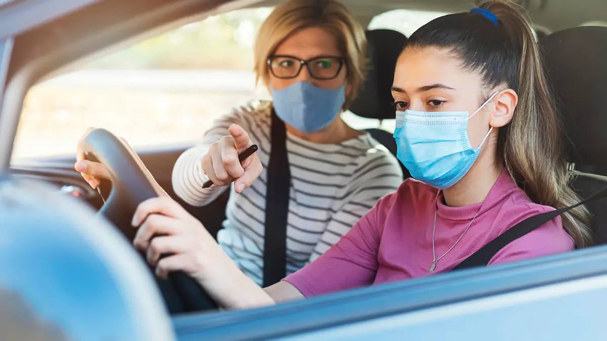A teenage girl with protective mask sitting behind the steering wheel of a car and listening to her mothers instructions as she drives.