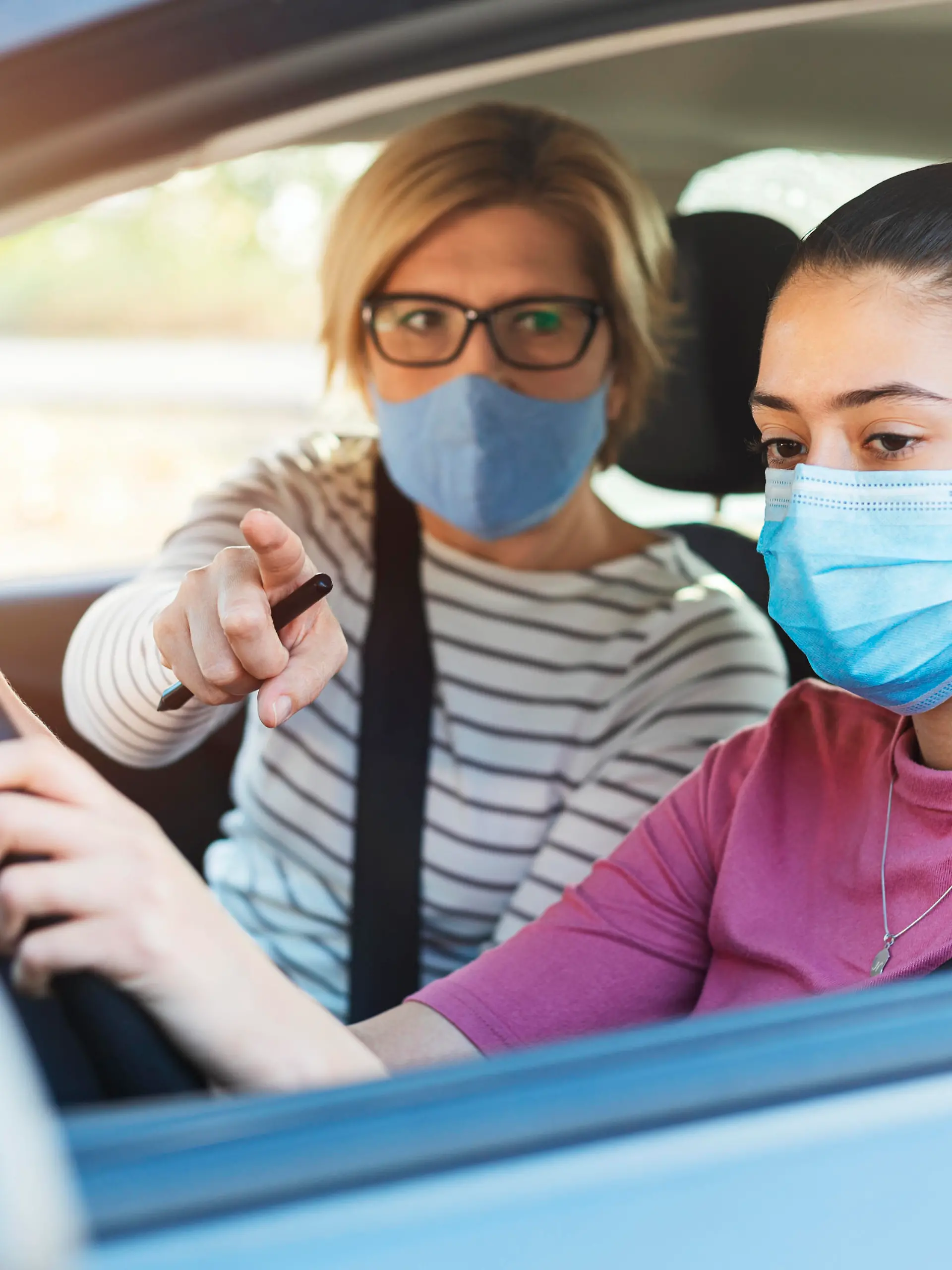 A teenage girl with protective mask sitting behind the steering wheel of a car and listening to her mothers instructions as she drives.