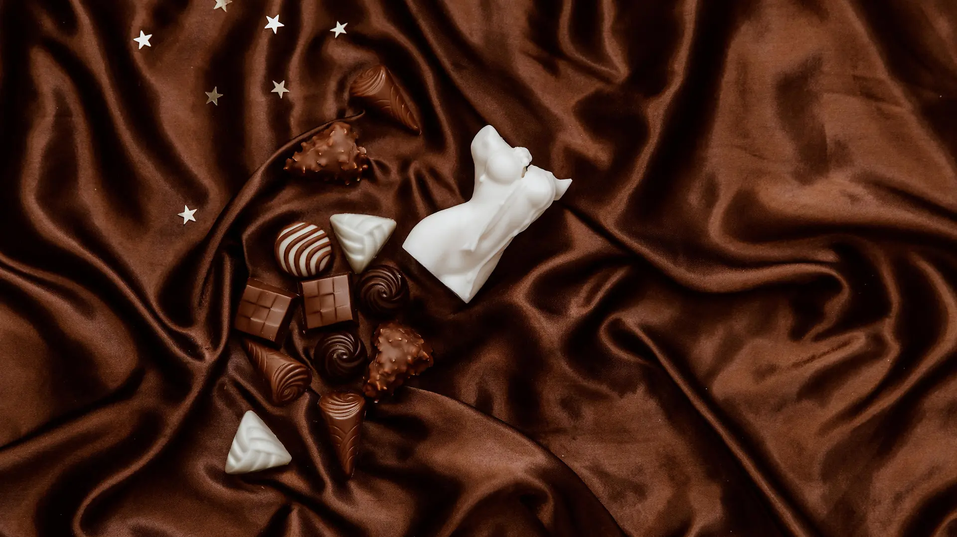 Styled flat lay of jewelry, chocolate candy, woman body shape candle on brown silky background. Lifestyle, beauty concept for blog, social media, magazine.