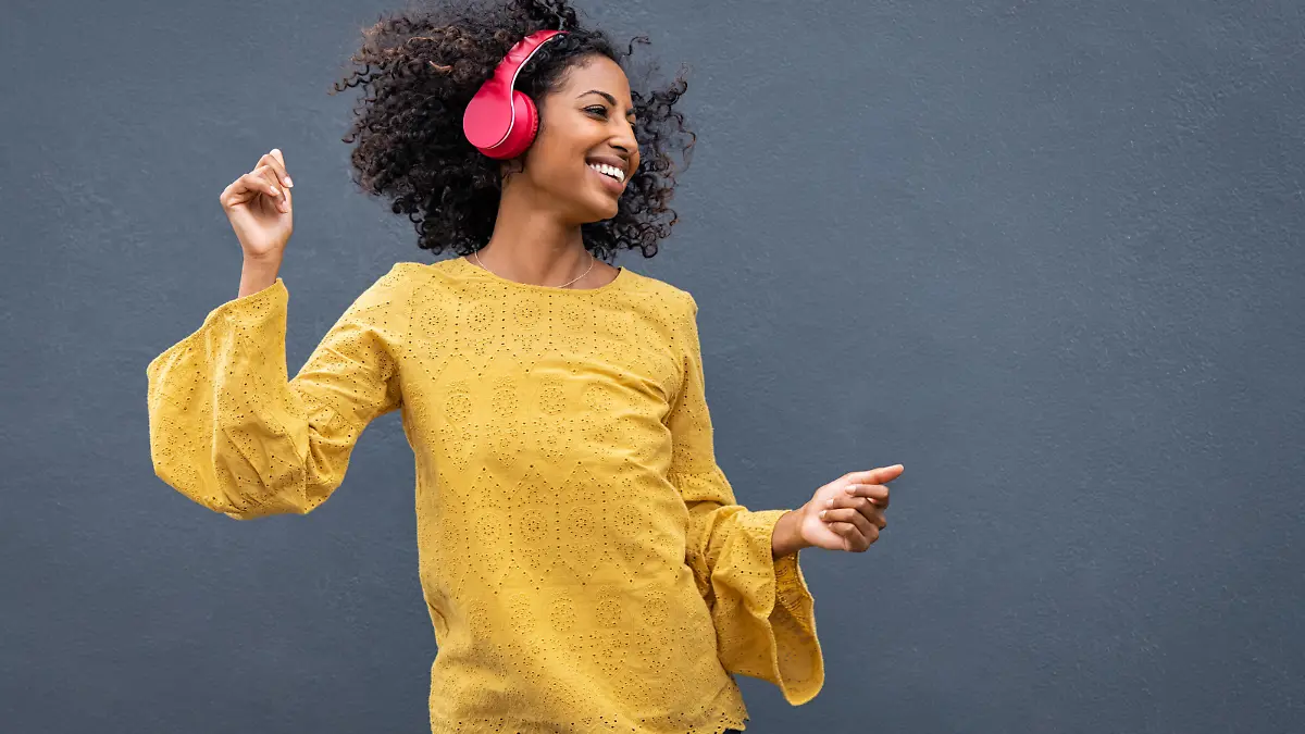 Beautiful black girl listening music in headphones from smart phone or mp3 player and dancing. Cheerful young woman with curly hair making dance moves to the rhythm of music while listening through wireless headphones isolated on grey wall.