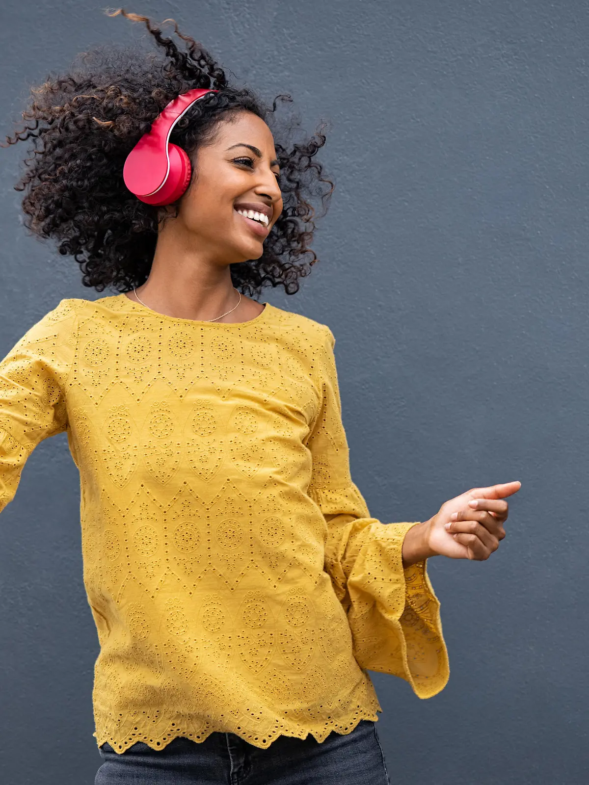 Beautiful black girl listening music in headphones from smart phone or mp3 player and dancing. Cheerful young woman with curly hair making dance moves to the rhythm of music while listening through wireless headphones isolated on grey wall.
