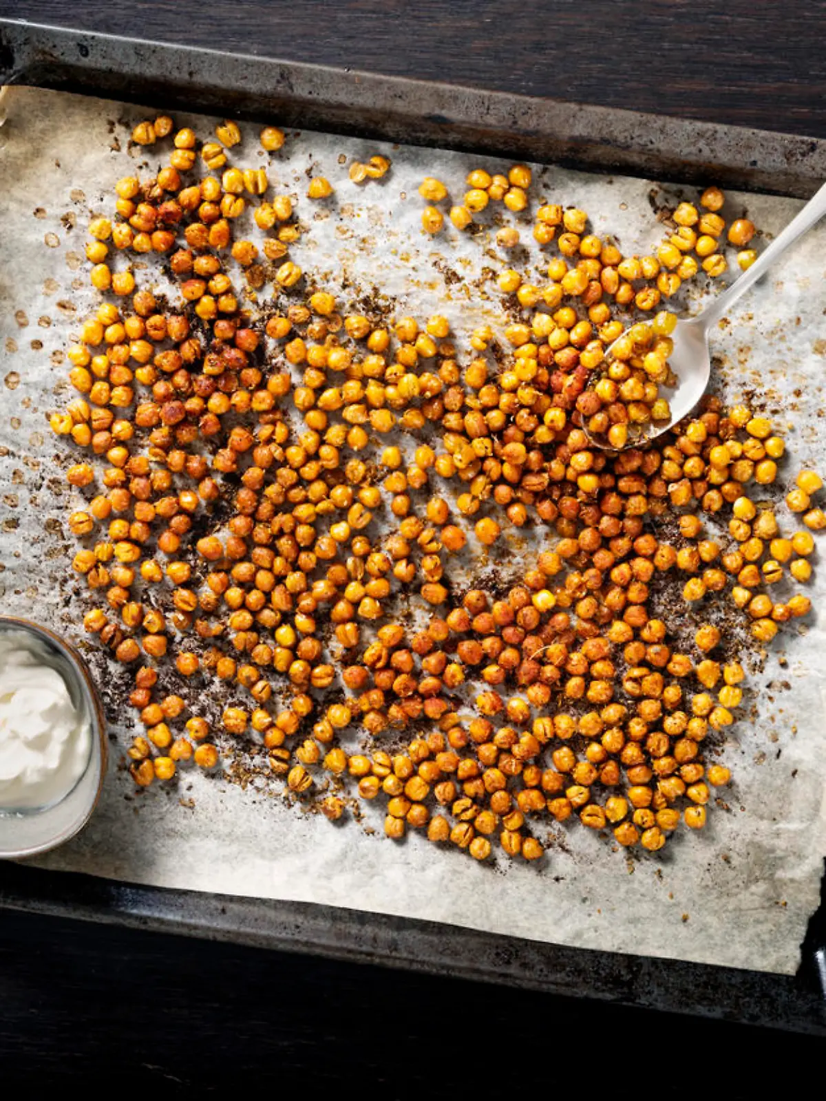 Tray of baked or roasted chick-peas seasoned with paprika and oregano. Overhead view looking down, photographed on a baking tray with baking parchment on it, colour, horizontal with some copy space. With a side bowl of soured cream or creme fraiche.