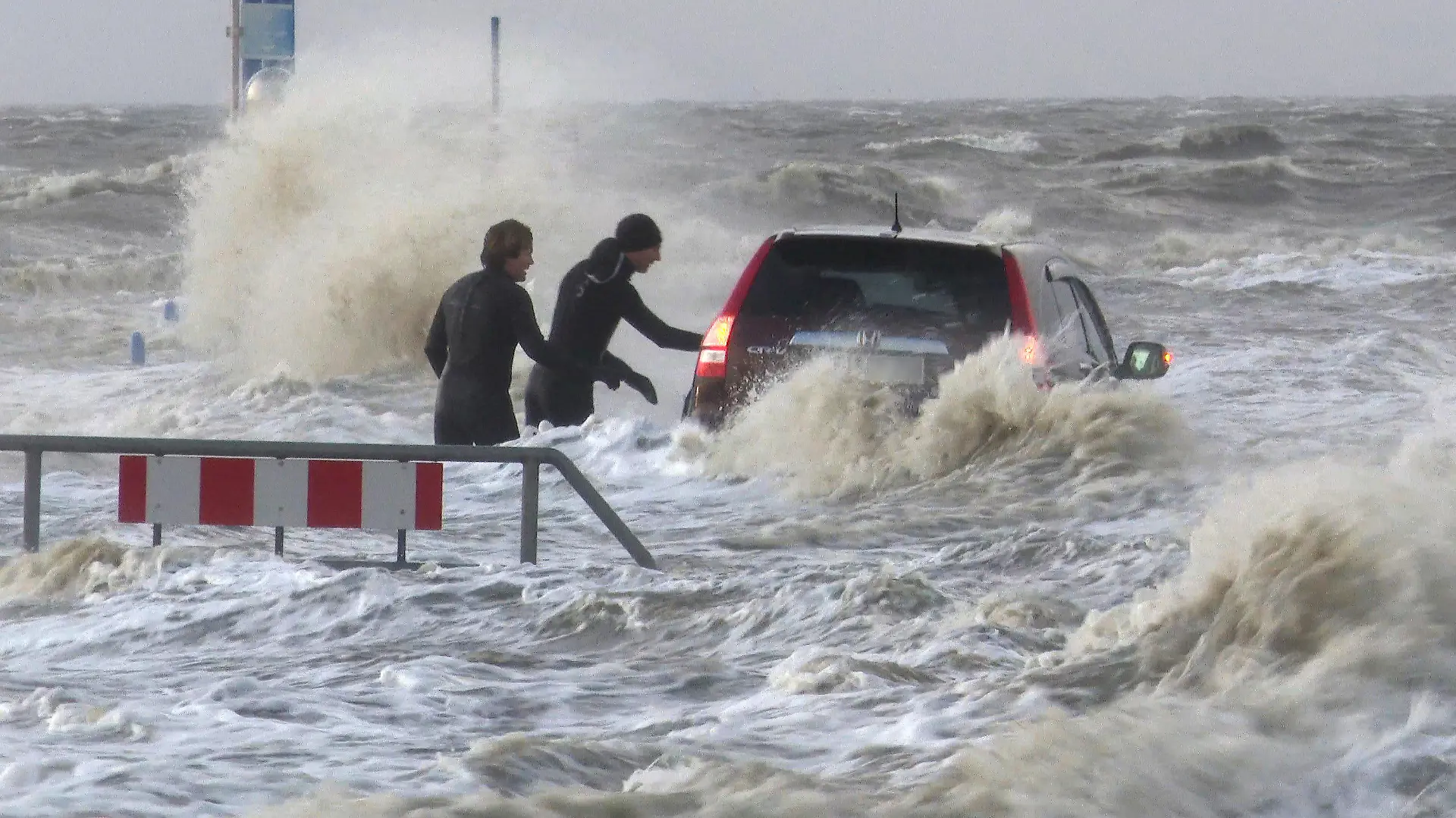 dpatopbilder - SCREENSHOT - 15.12.2019, Schleswig-Holstein, Dagebüll: Ein Auto steht auf einem Parkplatz der Mole des Fähranlegers am Hafen von Dagebüll (Schleswig-Holstein) im Wasser der Nordsee. Zwei Kitesurfer schauen in das Fahrzeug eines Unbekannten. An der Nordseeküste gab es am Nachmittag eine Sturmflut. (Screenshot, bestmögliche Qualität) Foto: Bodo Marks/dpa - ACHTUNG: Autokennzeichen gepixelt +++ dpa-Bildfunk +++