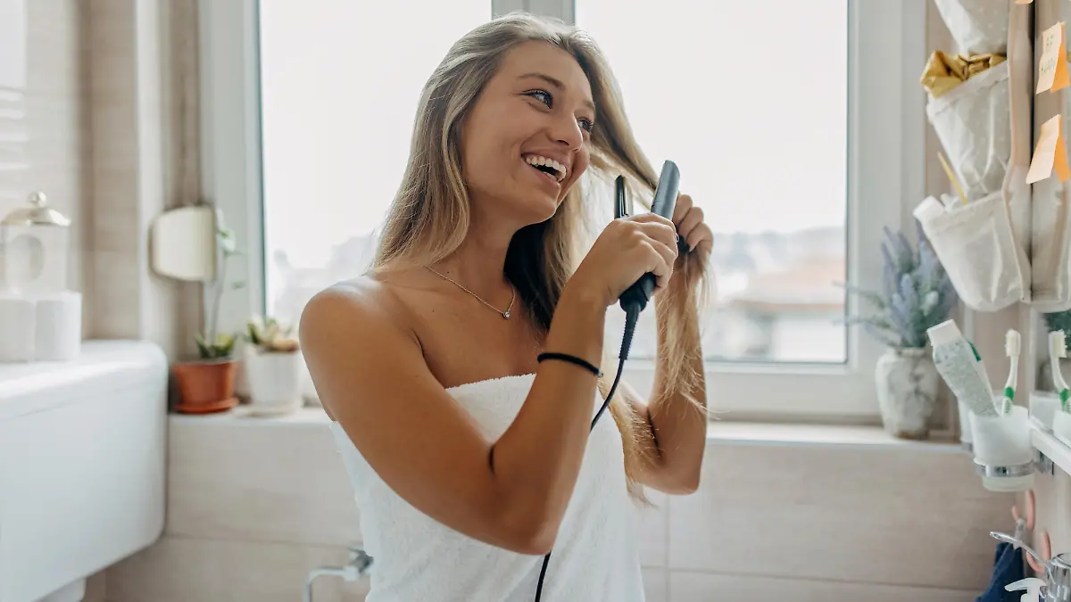 A young Caucasian woman is using a hair straightener while looking at the mirror with a smile on her face.