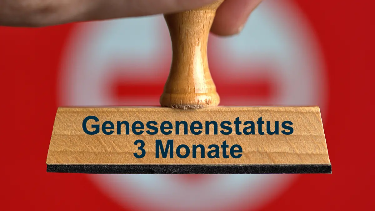 [M] Ein symbolischer Holzstempel mit der Aufschrift "Genesenenstatus 3 Monate", gehalten von einer Hand im Anschnitt vor einem unscharfen Hintergrund mit einem roten Kreuz. Foto mit Composing als Symbolbild zur Verringerung der Dauer, die eine mit Corona infizierte Person nach überstandenen Infektion als genesen gilt. Diese Dauer wird aufgrund der ansteckenderen Omikron-Variante von 6 auf 3 Monate verkürzt. || Modellfreigabe vorhanden