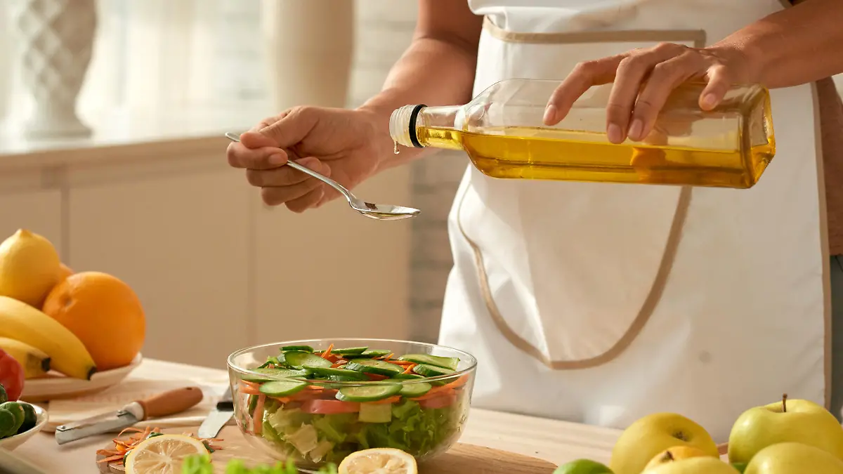 Female food blogger wearing apron pouring olive oil into vegetable salad while standing at modern kitchen, close-up shot