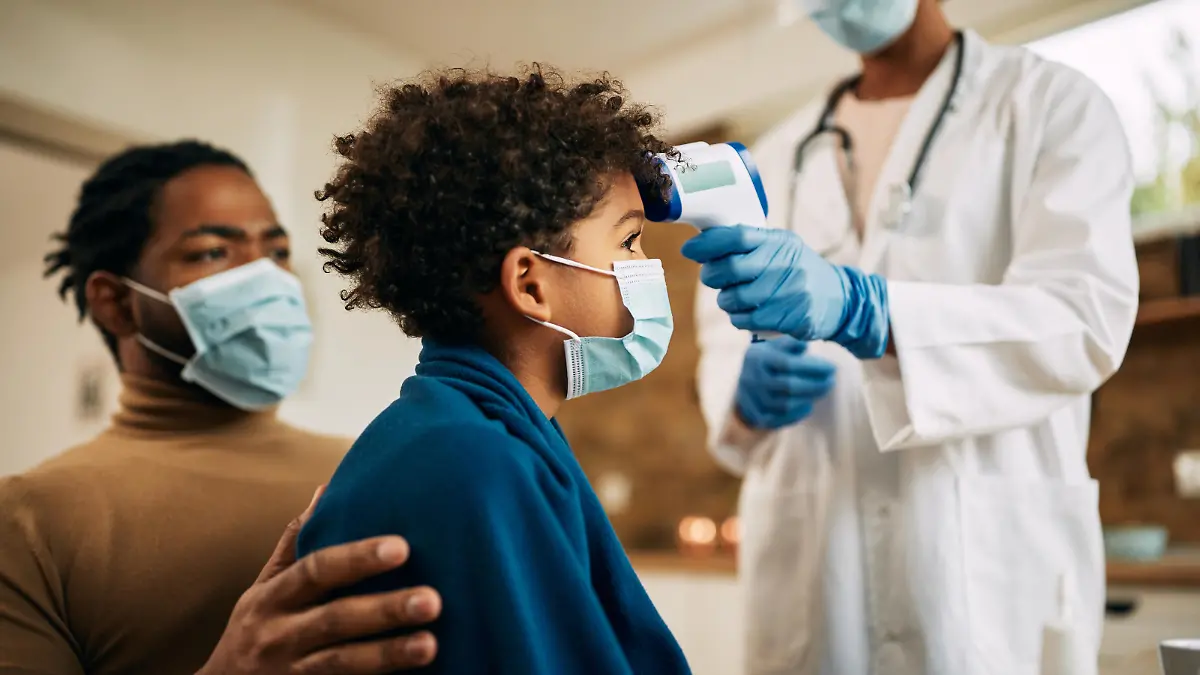 Close-up of pediatrician using infrared thermometer and measuring boy's temperature at home during coronavirus pandemic. Focus is on black boy.